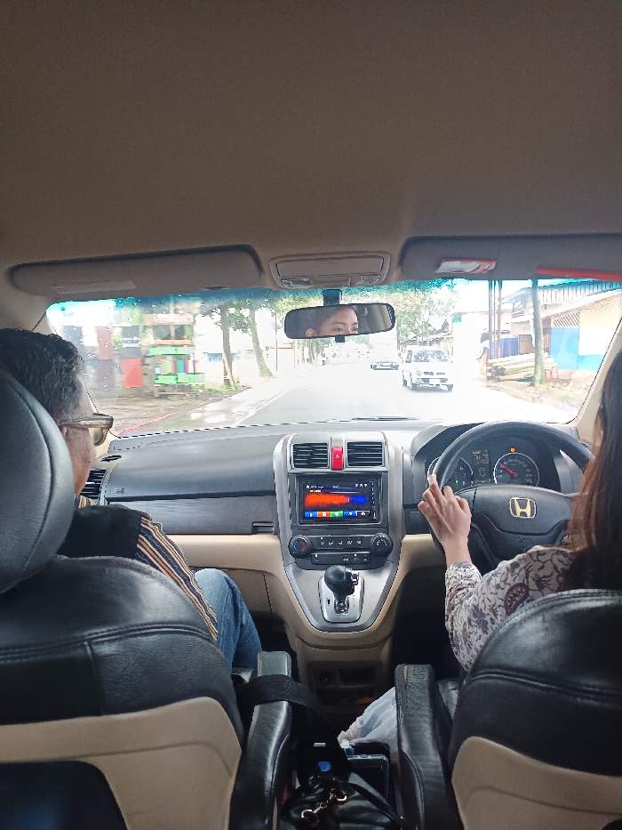 The image depicts the interior of a Honda CR-V, viewed from a rear passenger seat looking forward through the windshield, on a road in Cikaroya, Indonesia.
The vehicle's interior features a black dashboard with a tan lower section. A touchscreen infotainment system on the center console displays a multi-colored spectrum. An automatic gear shifter is positioned below it. The driver, a female, has her right hand on the steering wheel, which bears a Honda emblem. Her face is visible in the reflection of the rearview mirror. A male passenger is seated in the front passenger seat, seen from behind; he wears glasses, a striped shirt, and blue jeans. In the area between the front seats, a black bag, a mobile phone, and a water bottle are visible.
Outside the vehicle, a paved, two-lane road appears damp. On the left side, a structure with red, green, and blue panels is visible, along with other buildings and trees. On the right, residential-style buildings and a stack of long wooden or bamboo poles are situated near the roadside. Several other vehicles, including a white minivan, are visible on the road ahead in the distance. The sky is overcast.