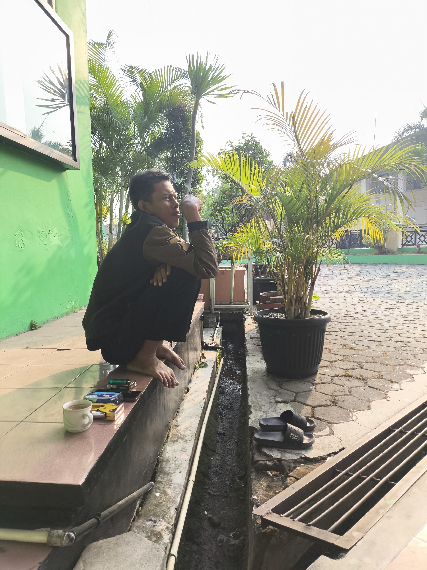 A man is squatting on a ledge outside a building. He is wearing a brown and black jacket and black pants. The building has a bright green wall and a large window with a reflection of palm trees. In front of the man, on the ledge, are a cup of coffee, several packs of cigarettes, a mobile phone, and a stapler. Behind him, to his right, is a large potted palm tree. A drainage ditch runs along the ground in front of the ledge. On the other side of the ditch, on the paved ground, are a pair of black slides with "SPECIAL FORCE" written on them. The setting appears to be an outdoor area in Cilegon, Indonesia, likely in a residential or commercial area given the building and paved surroundings. The sun is shining, casting shadows, indicating it is daytime.