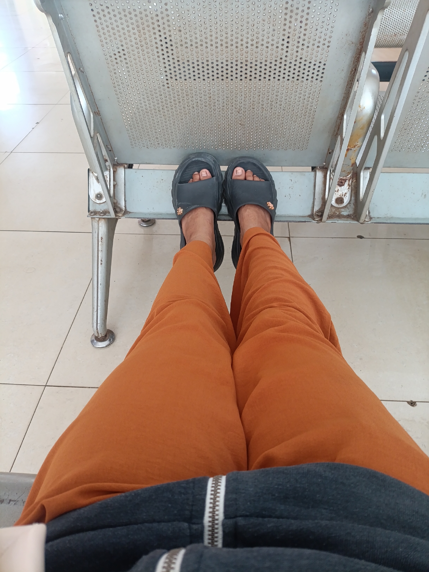 A first-person perspective shot shows a pair of feet wearing black slide sandals, clad in rust-orange trousers and a dark grey hooded jacket. The feet are resting on a light-colored tiled floor. Behind the feet, a metallic bench with a perforated seat is visible, likely in a public waiting area. The bench shows signs of wear and tear, with some rust visible. The overall scene suggests a casual setting, possibly a waiting room or public space in Jonggol, Indonesia.