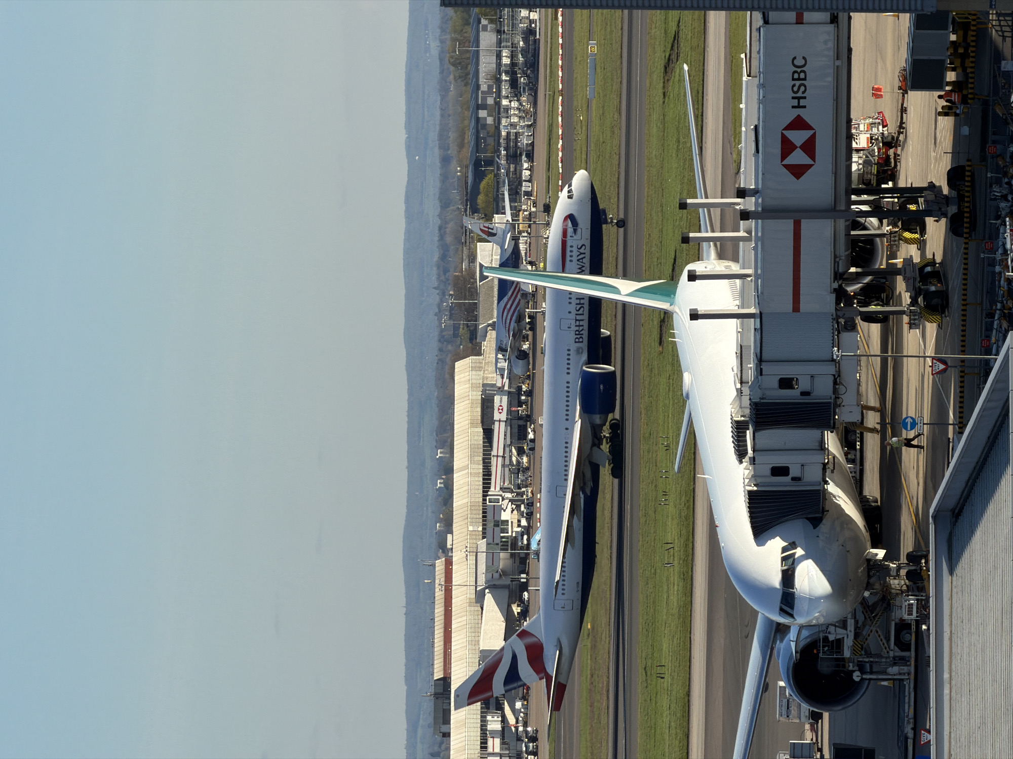 The image shows an aerial view of planes at Heathrow Airport, London, United Kingdom. A British Airways plane is docked at a terminal, with an HSBC-branded jet bridge connected. Another British Airways plane is on the runway nearby. Other planes are also visible in the distance, alongside airport buildings and ground vehicles. The sky is clear, and there is a general sense of daytime activity at a major international airport.