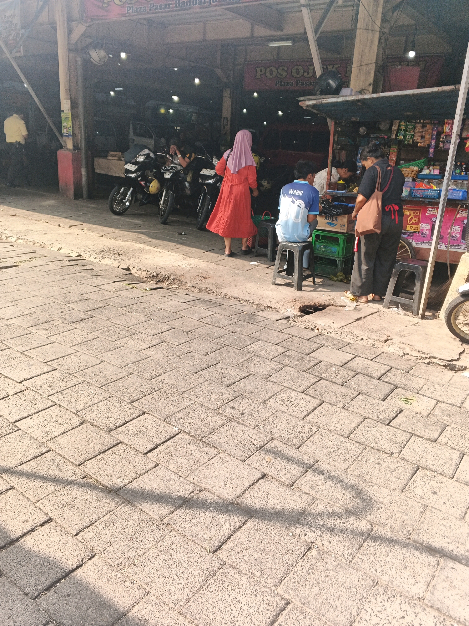 The image captures a street scene in Ungaran, Indonesia, likely a commercial area. In the background, a building with a sign reading "POS OJ" is visible. To the left, several motorcycles are parked. In the foreground and middle ground, people are present. A person in a red dress and a pink hijab stands near the motorcycles. Two individuals are seated on stools: one wearing a blue t-shirt with "WAHID" printed on it, and another person whose face is obscured. A third person, wearing dark clothing and carrying a brown bag, stands near a stall selling various items. The ground is paved with interlocking bricks, with some areas showing damage or wear. The lighting suggests it is daytime, with shadows cast on the pavement.