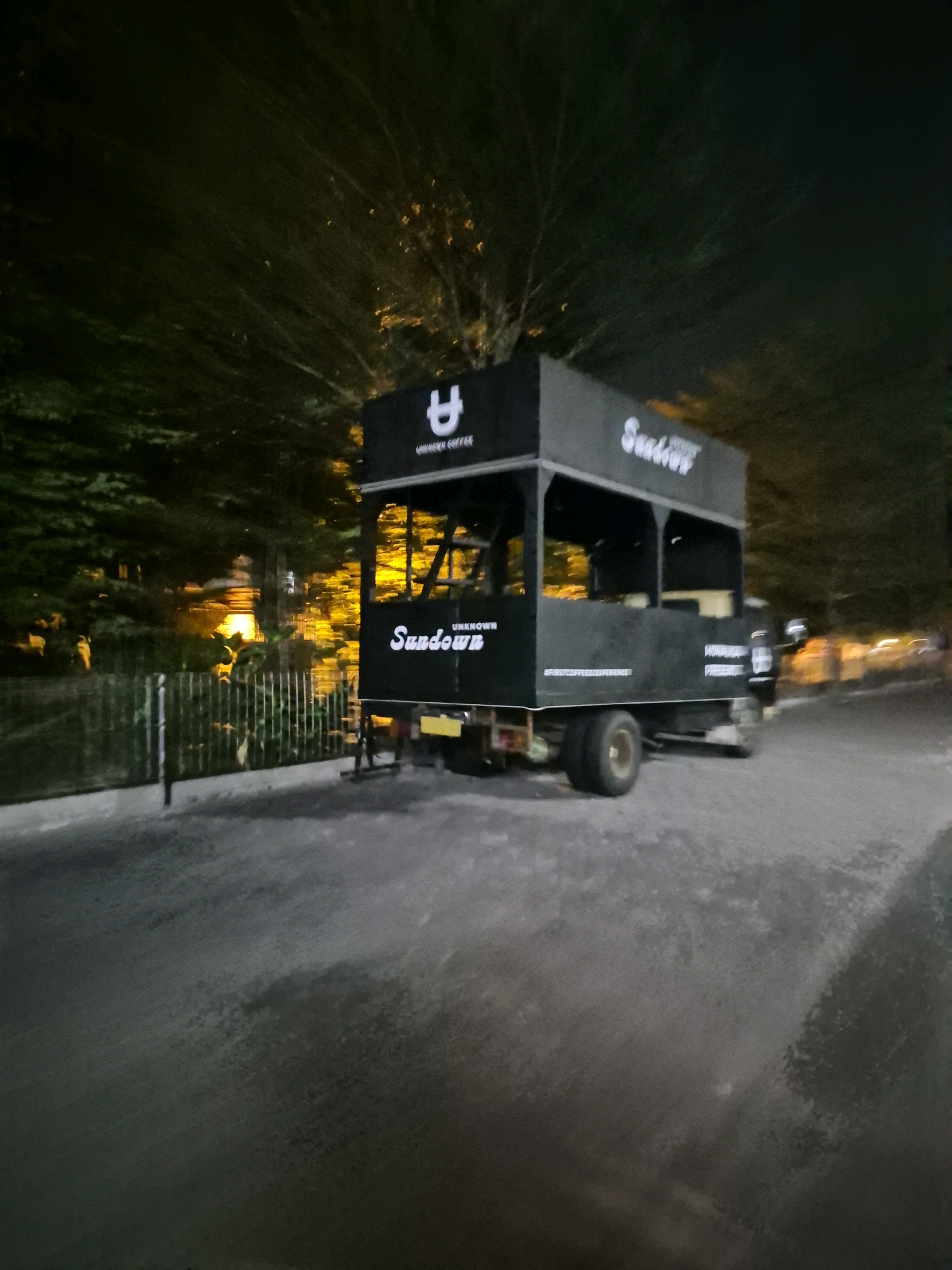 A dark, open-sided truck with a black canopy is parked on an asphalt road at night. The truck has the word "SUNDOWN" printed in white on its side, along with other text that appears to be branding. A logo consisting of a stylized "U" is also visible on the canopy. The truck is illuminated by streetlights and the headlights of passing vehicles, creating bright streaks of light in the background. Trees with dense foliage line the street, and a metal fence is visible to the left of the truck. The overall scene suggests a mobile coffee shop or food truck operating in a semi-urban or suburban area at night. The location is identified as Baturan, Indonesia.