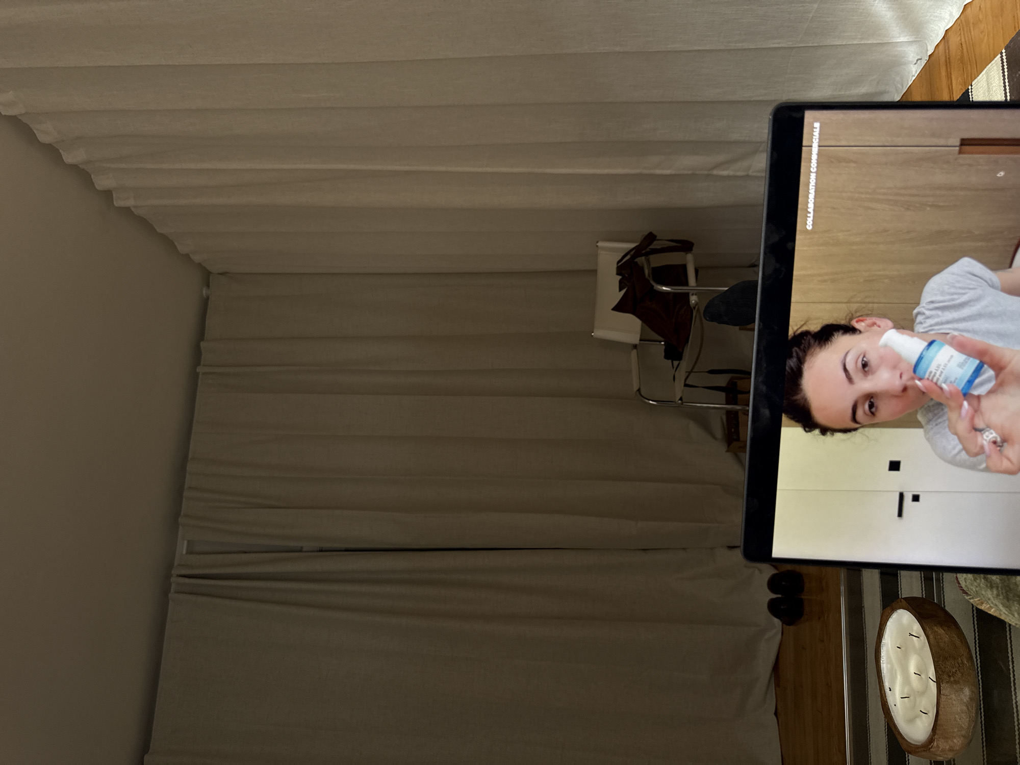 The image captures an indoor scene, likely a home in Montreal, Canada, illuminated by soft, artificial light, suggesting evening. The dominant feature is a large digital screen, possibly a tablet or monitor, placed on the right side of the frame. The screen displays a woman with dark hair pulled back, wearing a light-colored top, holding a small blue and white bottle, resembling a cosmetic product. She is looking towards the viewer. Text visible on the screen's display reads "COLLABORATION COMMERCIALE" in the upper left corner.
The room's decor includes light beige pleated curtains or blinds covering the left wall, extending across much of the background. In the foreground, below the screen, a striped rug is visible, upon which rests a decorative wooden bowl filled with multiple white candles. To the left of the screen, on a wooden floor, a pair of black slippers or shoes are present. Further back in the room, a white chair with metal accents, and a dark piece of fabric or bag draped over it, is partially visible. The overall impression is that of a quiet, comfortably lit domestic setting.