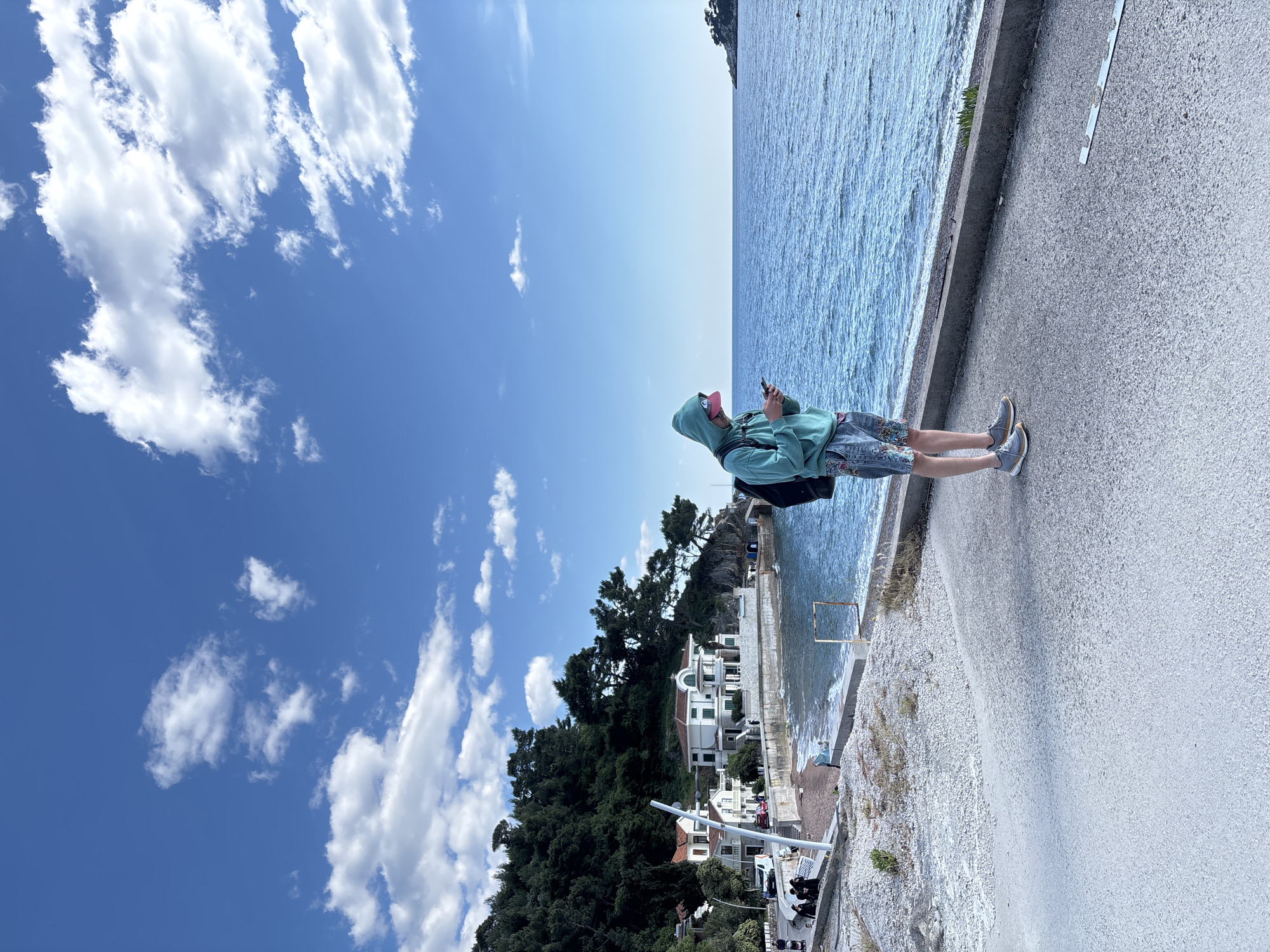 The image displays a bright, sunny coastal landscape with a person standing on a paved pathway by the sea. The image is oriented such that the person is standing upright, with the sea to their right and a hillside to their left.
The central figure is a person wearing a light green or teal hooded sweatshirt, patterned shorts (appearing to have floral or abstract designs), a pink baseball cap worn backward, and grey sneakers. They have a dark backpack on and are holding a dark smartphone with both hands, seemingly engaged with the device.
To the right of the person, the blue-green sea stretches into the distance, with gentle ripples and small waves visible near the shore. A concrete barrier or wall separates the paved walkway from the water. In the far distance, a headland or piece of land extends into the sea.
To the left of the pathway, there is a slope covered with gravel or small rocks. Further back, a dense array of dark green trees, possibly pines or other conifers, covers a hillside. Nestled among these trees and closer to the water's edge are several white buildings with distinct architectural styles, including one with prominent arched features and multiple levels, suggesting a Mediterranean or European coastal setting.
The sky above is a vivid blue, adorned with numerous white, fluffy clouds of various sizes and shapes, indicating clear weather. The strong, even lighting suggests it is daytime, likely mid-morning or afternoon. There is no visible text in the image.