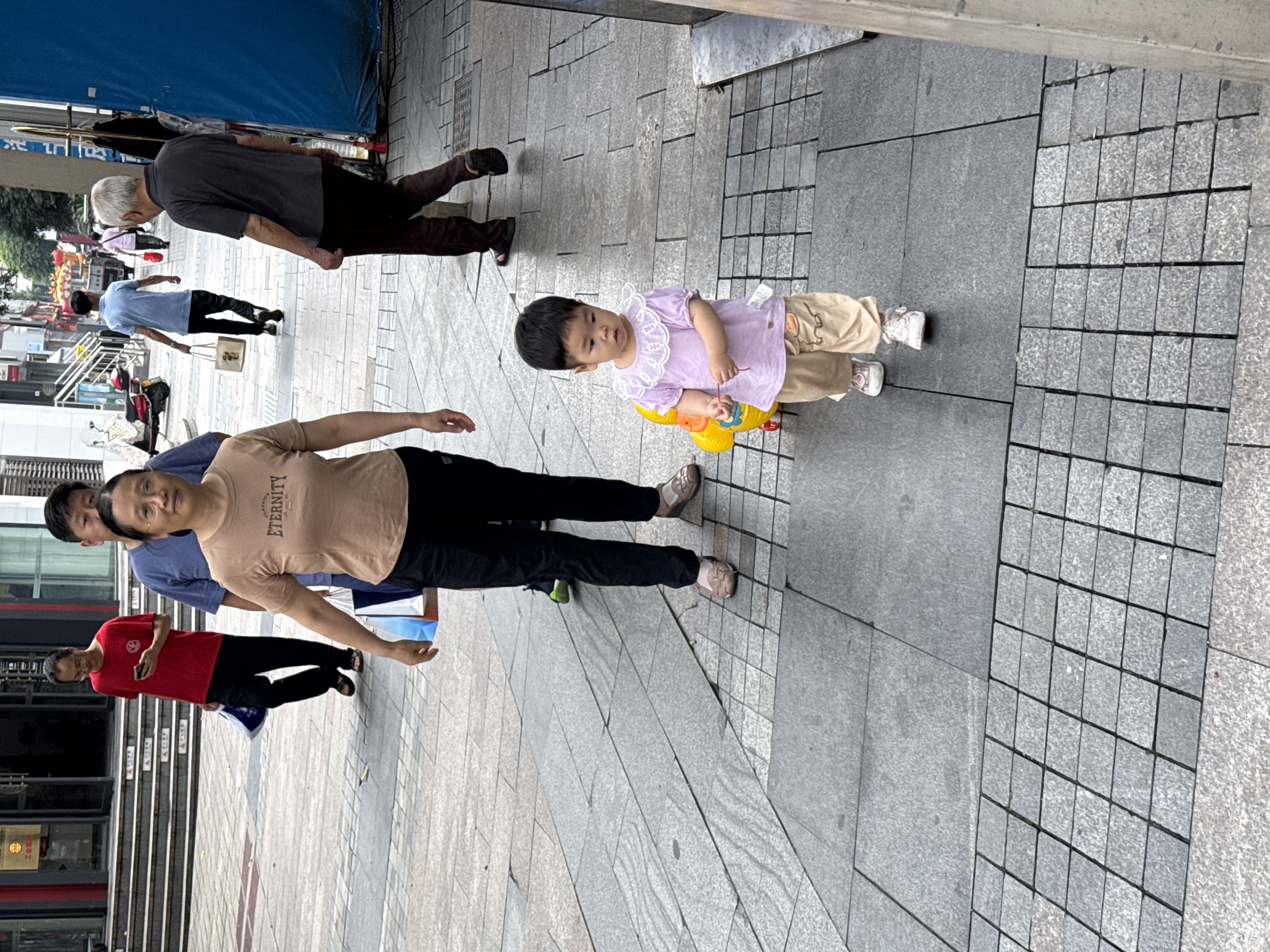 The image shows a street scene in Jiangjin, China. Several people are walking on a paved sidewalk. In the foreground, a woman in a tan t-shirt and black pants walks with her arms outstretched. Behind her, a man in a blue t-shirt and black pants walks with a shopping bag. To the right of the woman, a toddler in a purple top and beige pants stands holding a yellow toy. Further back, an older man in a grey t-shirt and dark pants walks with his arms down. The scene appears to be daytime, with bright lighting. The background shows buildings and a blue tarp, suggesting an urban environment. The people's expressions are neutral. Text is visible on the woman's t-shirt, reading "ETERNITY".