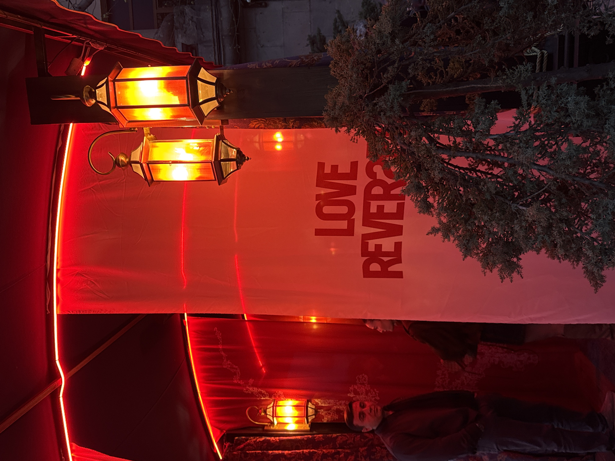 The image depicts an outdoor or semi-enclosed area illuminated primarily by red light, located in the city of Yerevan, Armenia. Overhead, red LED strip lights trace structural elements and the perimeter of red fabric awnings or coverings. A tall white banner or panel, vertically oriented, displays the partial red text "LOVE REVER?" on its lower section. To the mid-right, two ornate, antique-style lanterns, emitting an amber-orange glow, are mounted on a dark, vertical wooden post. Another similar lantern is visible on the far left. A dense evergreen shrub or tree occupies the lower right and mid-right foreground, with dark green foliage. Two individuals are present: a man in a dark top stands on the left, facing generally forward, under intense red lighting. Partially visible behind the white banner on the right is another individual's head and upper torso, facing left, also illuminated by red light. The environment outside the immediate red-lit area appears dark, with indistinct architectural features visible in the upper right background.