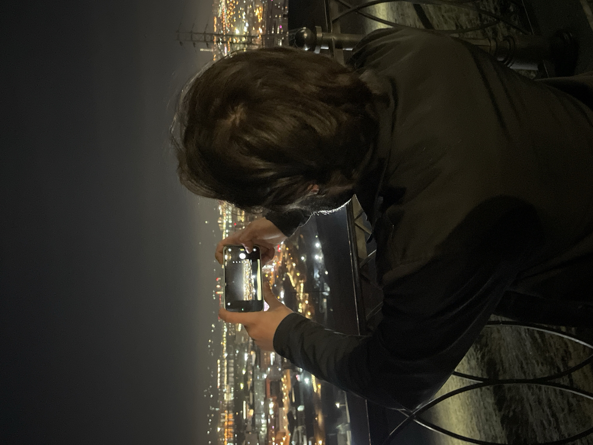 The image captures a nighttime view of a vast cityscape from an elevated perspective, likely an observation deck or tall building, in Krasnoyarsk, Russia. The dominant subject is a person, seen from behind, with dark hair and wearing a dark, long-sleeved garment, possibly a jacket.
The individual is actively engaged in photographing or videoing the illuminated urban landscape below using a smartphone, held horizontally with both hands. The screen of the smartphone reflects the bright, scattered city lights, suggesting it is capturing the same impressive view. A dark metal railing, possibly part of a balcony or barrier, is visible in the foreground, indicating the vantage point.
The background is dominated by the shimmering lights of the city, which stretch far into the distance and appear slightly blurred, creating a sense of depth and scale. The sky above the city is dark, contrasting sharply with the vibrant urban glow. The atmosphere appears somewhat hazy or misty, which diffuses the lights and adds to the visual effect. The time of day is clearly night. There is no discernible text visible in the image.