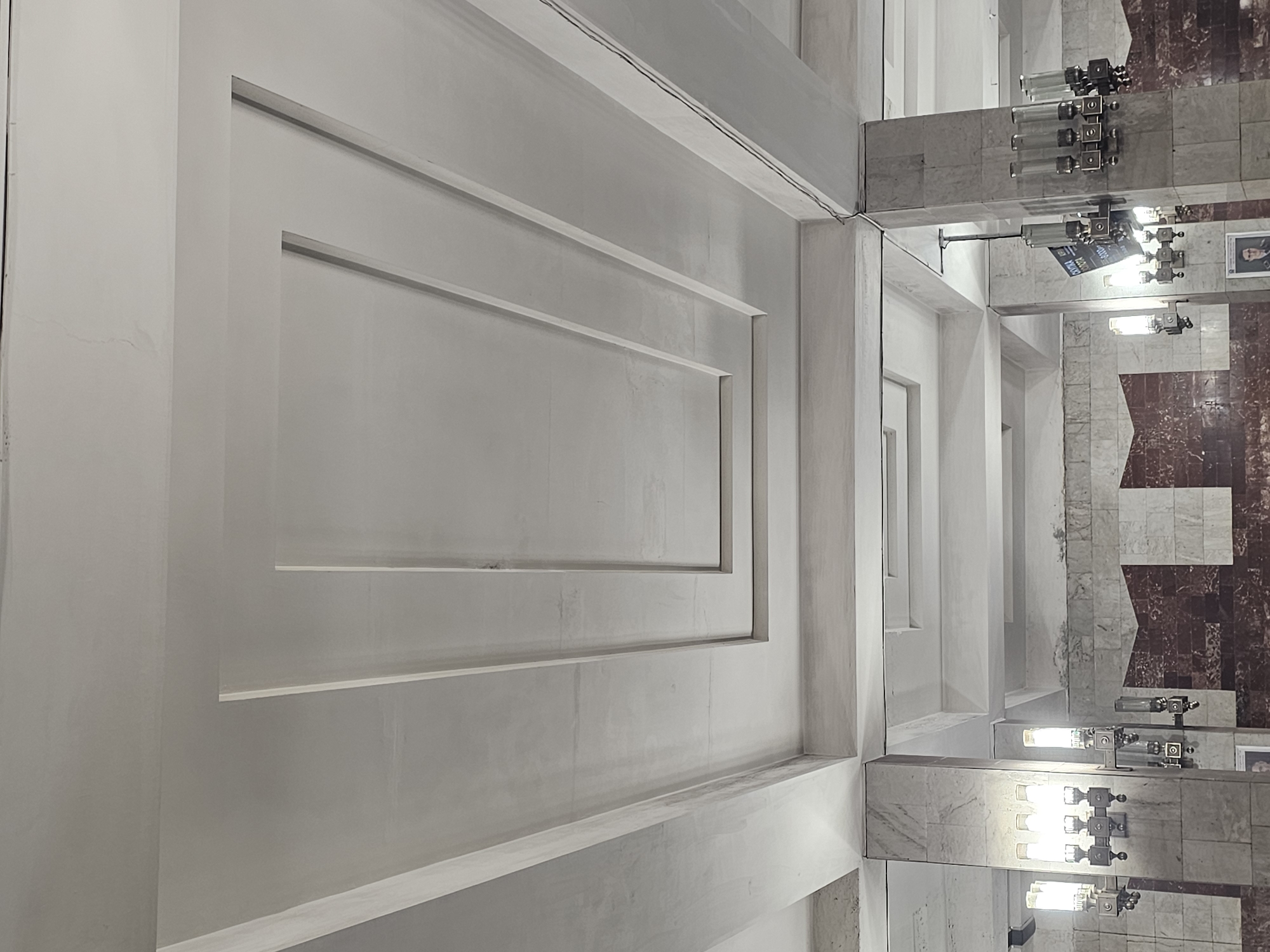 The image shows the interior of a building, likely a public space such as a metro station or an old administrative building. The ceiling features a layered, geometric design with recessed rectangular panels. The walls are clad in marble with a patterned section of darker red and white stone. There are multiple vintage light fixtures mounted on columns, some appearing as sconces and others as chandeliers. A digital display board is visible hanging from a column. The overall aesthetic suggests a retro or classical architectural style. The location is the metro station "Gorkovskaya" in Nizhny Novgorod, Russia. There are no people clearly visible in the frame.