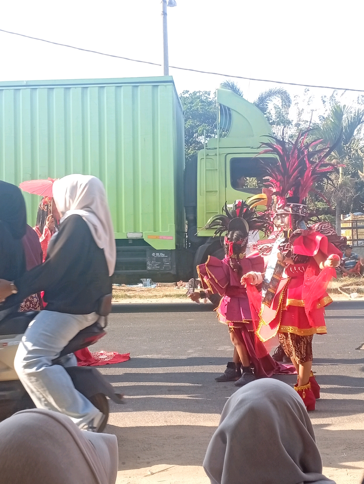 The image captures a street scene in Bancar, Indonesia, during what appears to be a parade or cultural event. In the foreground, a person wearing a light gray hijab is seated on a motorcycle, their legs clad in light gray pants. Beside them, another person in a black top and light gray pants is also on the motorcycle, their left hand resting on the handlebars. To the left, a person in a black hijab and a red garment is partially visible.
The central focus of the image is on two individuals in vibrant, traditional costumes. Both are adorned with elaborate feathered headdresses in red and black. The individual on the left wears a magenta top and skirt, with layered ruffles and gold trim. The individual on the right is dressed in a similar style but in a bright red outfit, also featuring gold embellishments and a flowing sash. Both are holding red fabric pieces.
In the background, a large, light green truck is parked, its ribbed metal side dominating the upper left portion of the frame. The truck appears to be a cargo truck. Behind the truck and the costumed individuals, palm trees and other foliage are visible, suggesting a tropical or subtropical environment. Further in the background, other people are seated on the ground, observing the event. The scene is set on a paved road under a bright, sunny sky.
