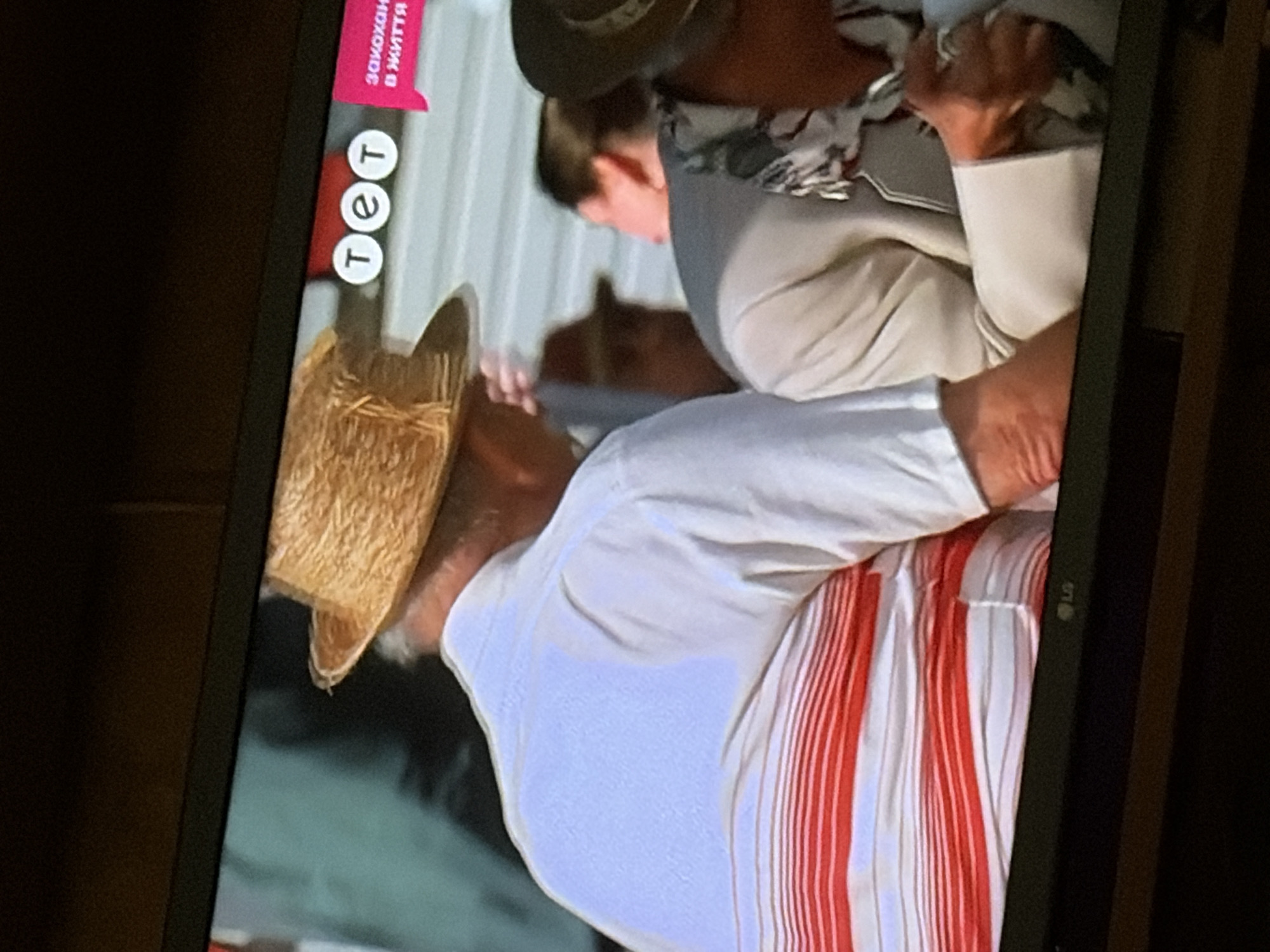 The image shows a close-up of a television screen displaying a scene with several people. In the foreground, a person wearing a straw hat and a white shirt with red stripes is visible from the back and side. To their right, another person is partially visible, wearing a light-colored jacket. A logo with three white circles containing the Cyrillic letters "т", "е", and "т" is displayed on the screen, along with pink text that appears to be in Cyrillic. The background is blurred and appears to be an outdoor setting. The bottom of the television screen shows the "LG" logo. There is no visual information in the image that specifically places the scene in the city of Osolynka, Ukraine.