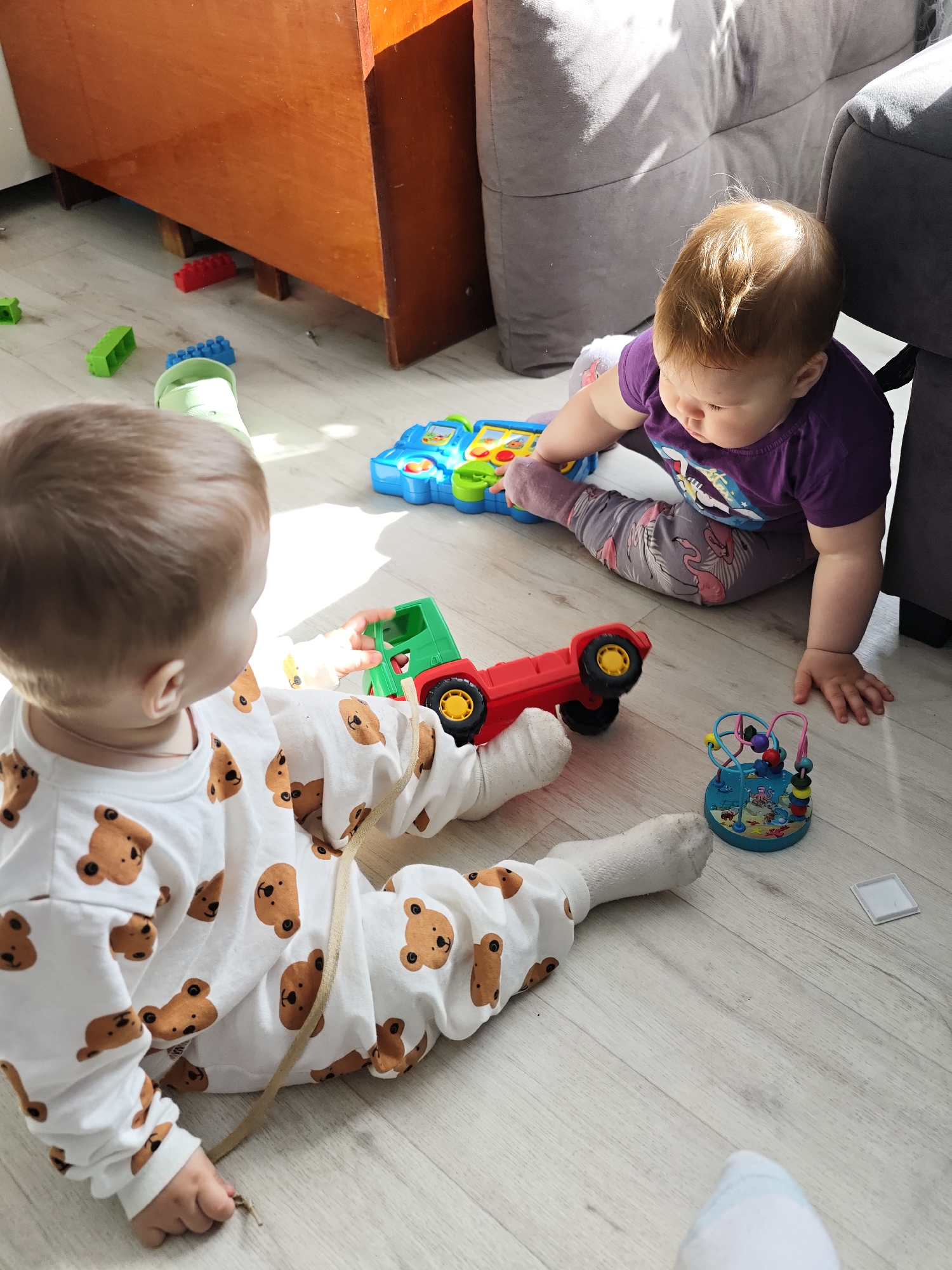 Two infants are playing on a light-colored wooden floor indoors. One infant, dressed in a white onesie with brown teddy bear prints, sits with legs extended and holds a green toy truck. The other infant, wearing a purple t-shirt and patterned leggings, sits near a gray couch and engages with a blue electronic toy. Additional toys are scattered on the floor, including a red toy truck and a blue bead maze. A wooden cabinet sits in the background, near the window which is the source of sunlight illuminating the scene. The location is Уральское, Russia.