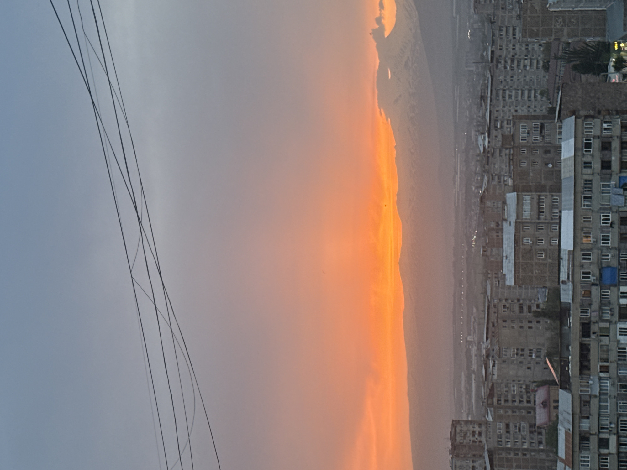 The image shows a view from an elevated position in Abovyan, Armenia, likely from a building or high point. The right side of the image depicts a dense urban landscape with numerous residential buildings, some appearing to be of Soviet-era construction. The buildings vary in height and architectural style. Windows are visible on the buildings, with some reflecting light.
To the left, the sky is dominated by a vibrant sunset, transitioning from a light gray to an orange hue near the horizon. The horizon line is punctuated by distant mountains. Power lines are visible in the upper left quadrant of the image.