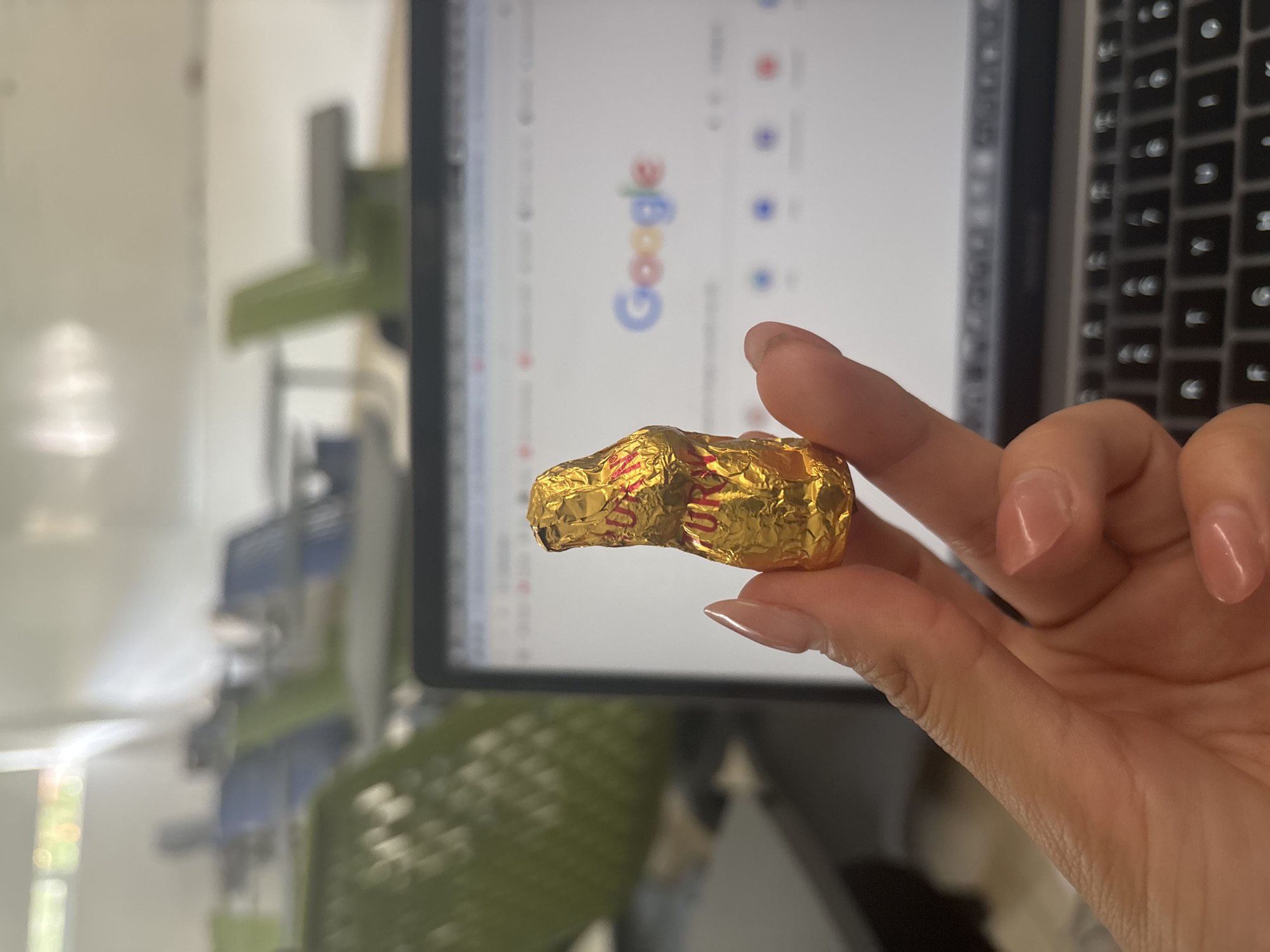 A person's hand holds a gold-wrapped chocolate candy in front of a laptop screen displaying the Google search engine homepage. The laptop is situated on a desk next to a keyboard. The background is a blurred office setting with a green filing cabinet. The location cannot be determined as Puebla, Mexico, from the information provided in the image.