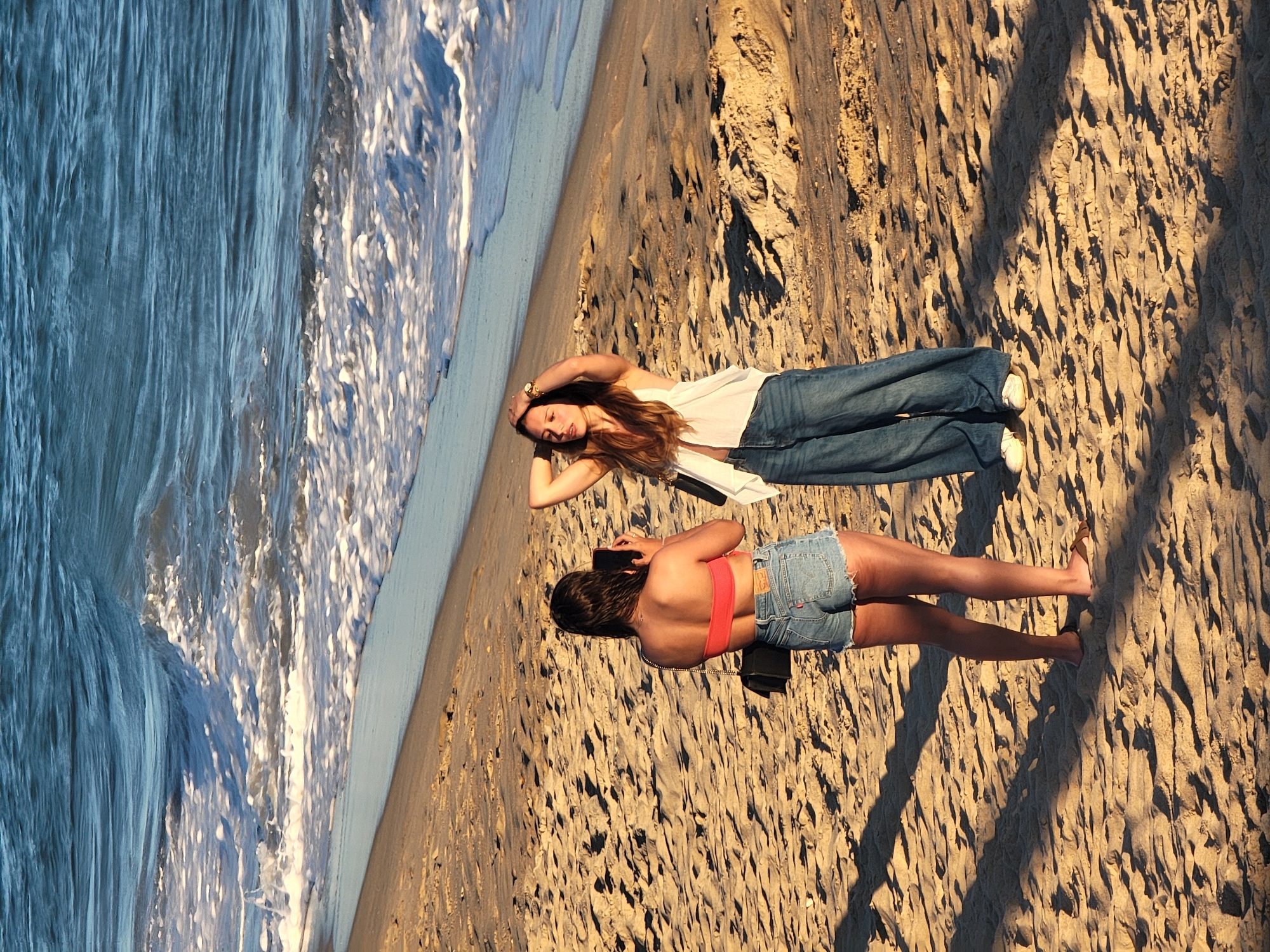 Two women are on a sandy beach near the ocean in Quarteira, Portugal. The woman on the left is facing away from the camera, holding a black smartphone and wearing a coral-colored bandeau top and denim shorts. She has a black purse slung over her shoulder. The woman on the right is facing towards the ocean and is angled towards the left. She has her hands on her head and is wearing a white sleeveless top and wide-leg blue jeans. She is also carrying a black purse. The ocean waves are visible in the background, and the sand is textured with shadows from unseen objects.