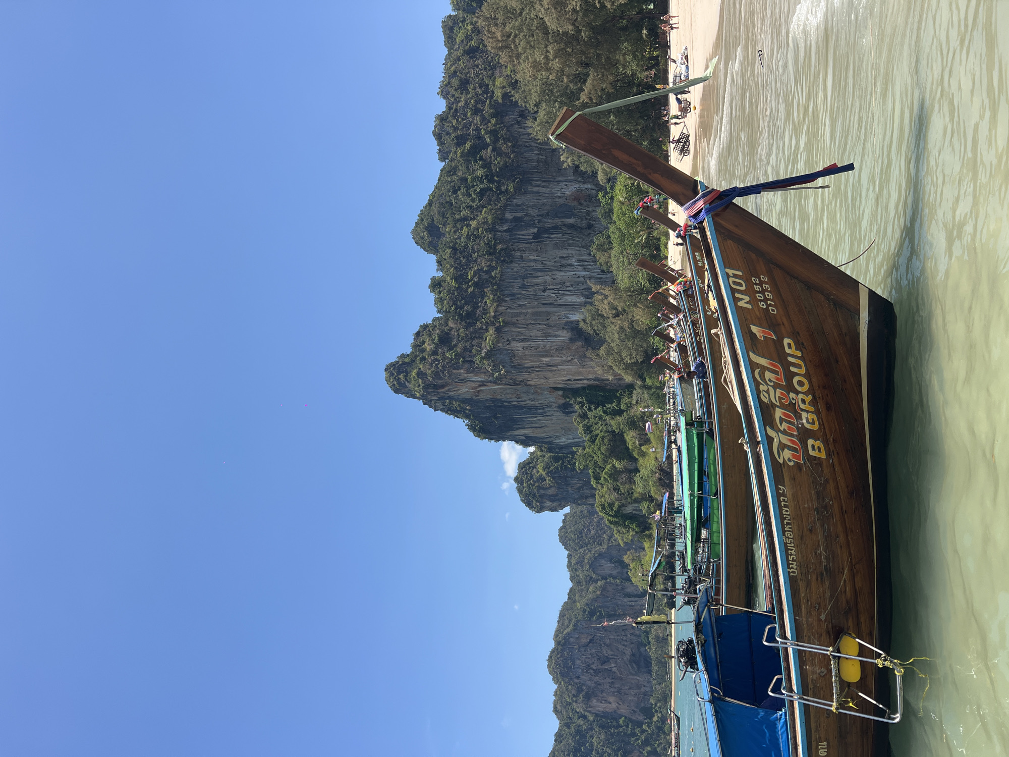 The image depicts a scene in Ban Ao Nang, Thailand. Several longtail boats are docked along a sandy beach with clear, light-colored water. The boats are wooden, with colorful accents and traditional adornments, including fabric tied to the prows. One boat has the inscription "B GROUP" in English, along with Thai script and the number "N01". Rocky cliffs covered in lush green vegetation rise in the background. The sky is a clear, bright blue. People are visible on the beach in the distance.