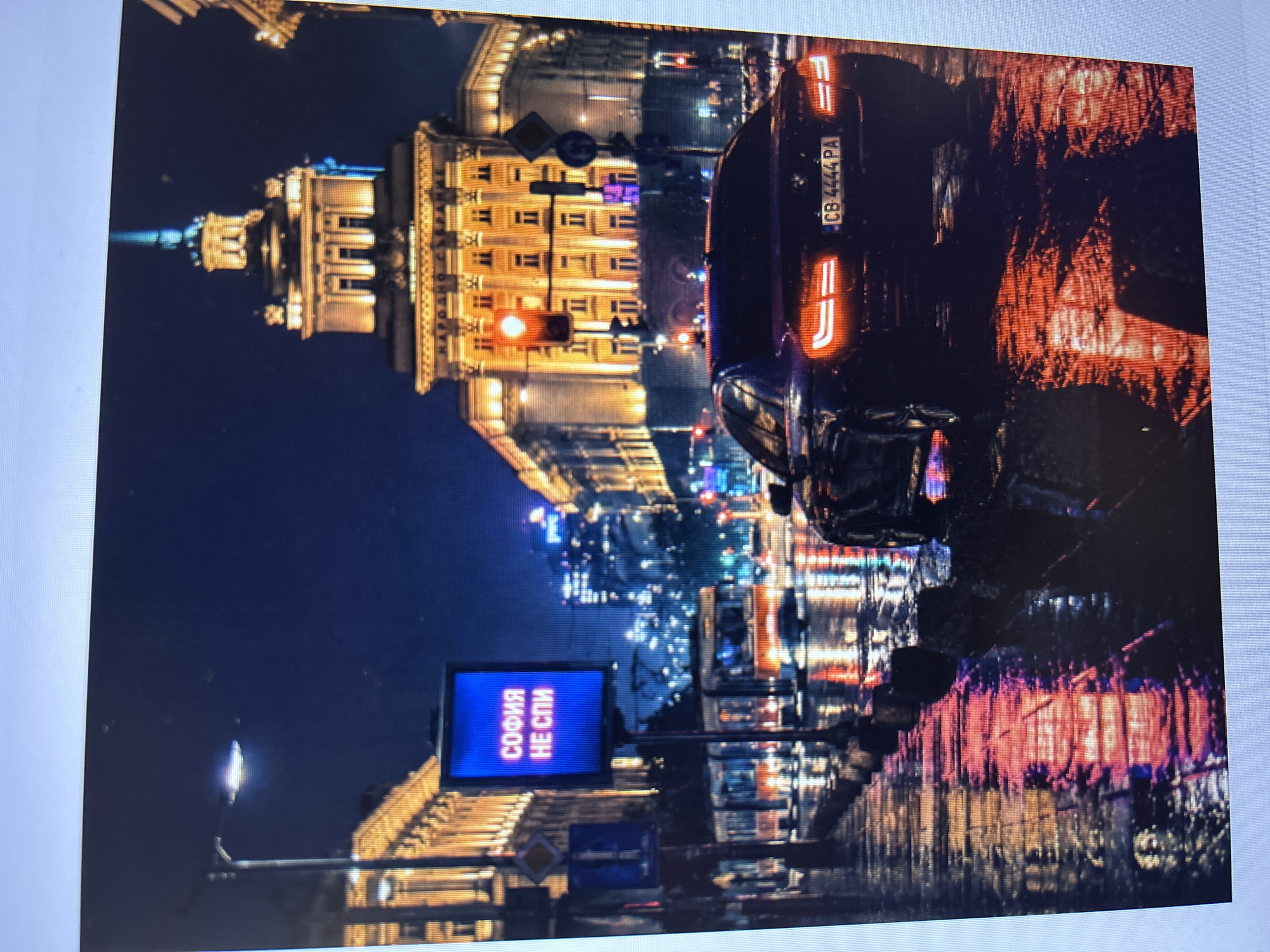 The image captures a nighttime scene in Sofia, Bulgaria, featuring a car stopped on a wet street, with reflections of city lights on the pavement. In the background, the illuminated architecture of the Largo, notably the former Party House, is visible. A blue digital billboard displays the phrase "Sofia does not sleep." The license plate "CB 4444 PA" is visible on the car. The scene suggests a rainy night in the city center.