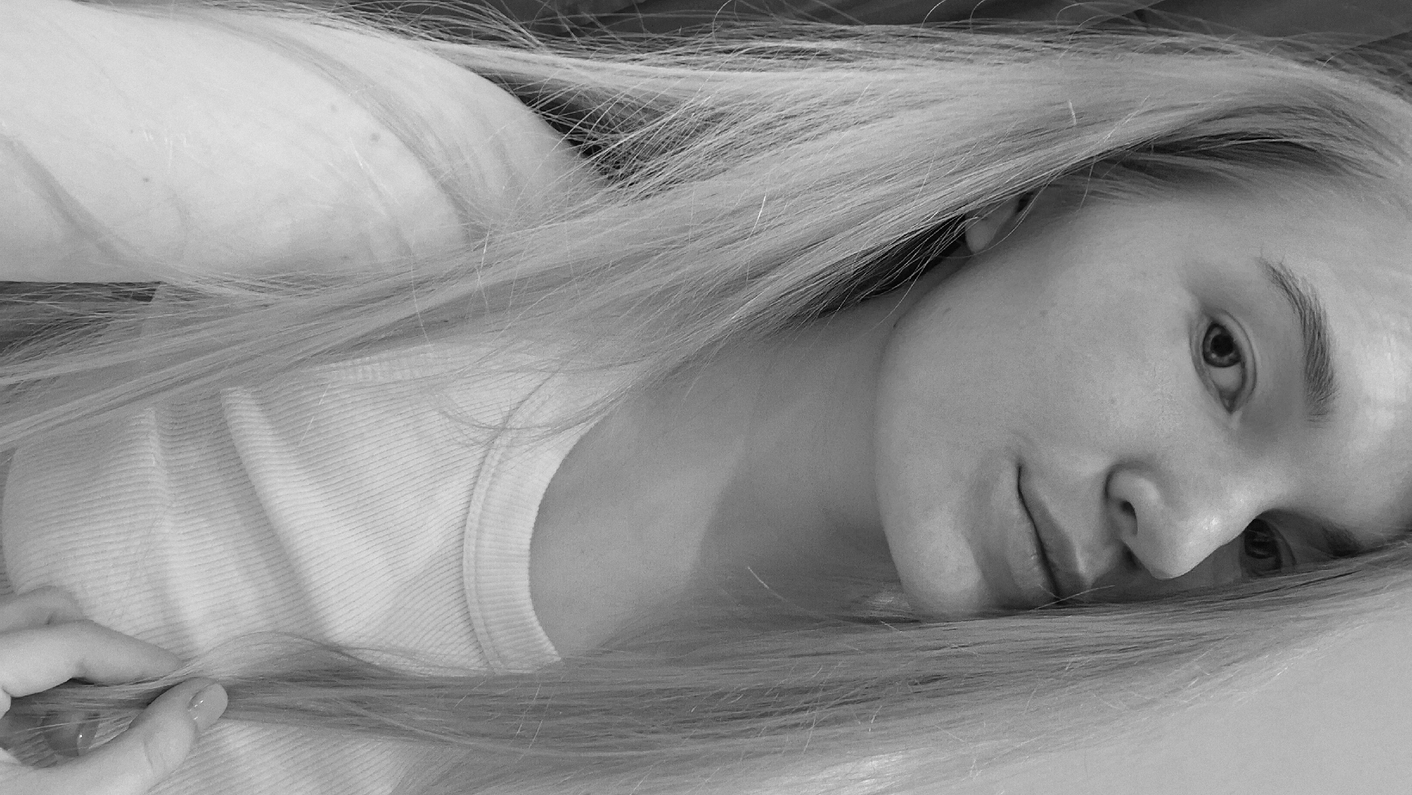 A monochrome, close-up, medium-shot photograph captures a young woman lying down, presumably indoors. Her face is turned towards the viewer, with a slight upward gaze. She has fair skin, defined eyebrows, and light-colored, possibly blonde, hair that is spread around her head and shoulder. The hair appears straight and fine-textured.
She is wearing a light-colored shirt, visible around her neck and shoulder area. Her left hand is partially visible in the lower left corner of the frame, gently holding a strand of hair. The background is blurry and indistinct. The location in the city of Unknown, Unknown is not discernible from the image.