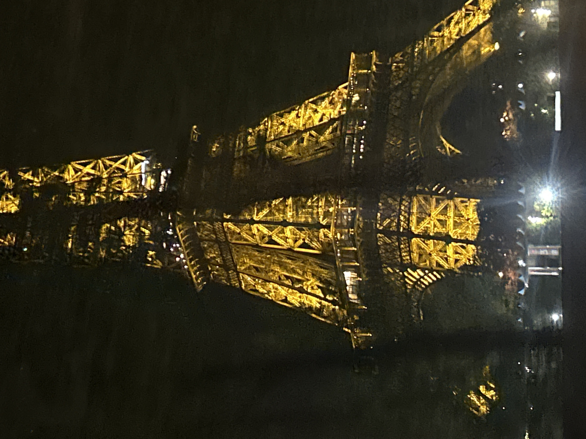 The image shows the Eiffel Tower in Paris, France, at night. The tower is illuminated with yellow lights and is reflected in a body of water, likely the Seine River. The sky is dark, and the surrounding area appears to be dimly lit.