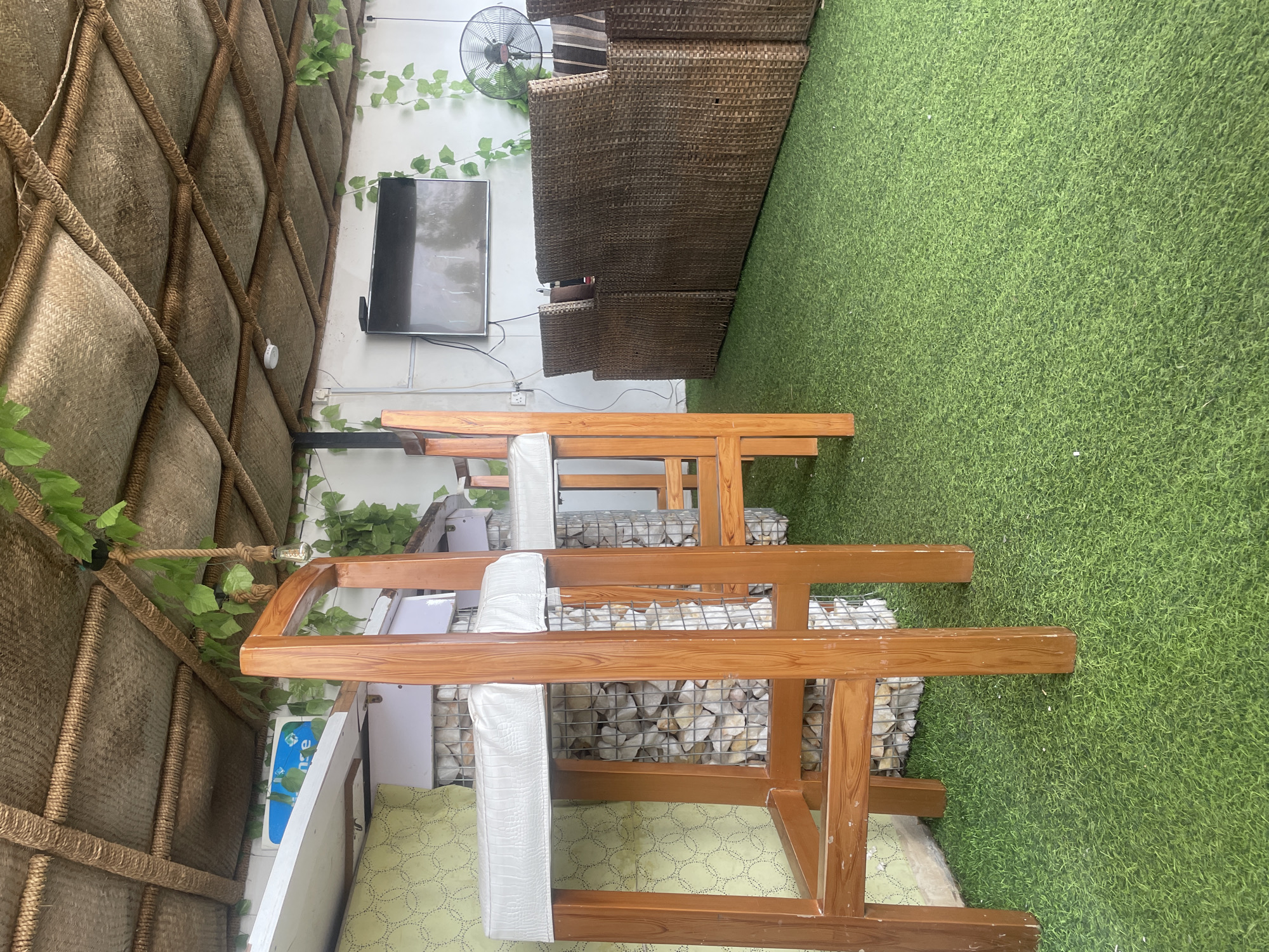 This media file depicts an outdoor seating area, likely in a warm climate. The scene is dominated by a vibrant green artificial turf floor, suggestive of a garden or patio setting. Several pieces of wooden furniture, possibly benches or tables, are arranged in the foreground. These are constructed from light-colored wood with visible grain, and some have white upholstered cushions. Behind the furniture, a wall is visible, decorated with a woven, textured material in a diamond pattern, interwoven with thick rope. Green artificial vines are draped over this wall and along the ceiling.
On the wall, a flat-screen television is mounted, displaying a reflective surface, suggesting it is either off or showing a dark image. A ceiling fan is also visible on the wall, along with some electrical outlets. Further back, there are stackable chairs made of dark, woven material, resembling rattan or wicker, adding to the outdoor lounge aesthetic. A metal cage filled with decorative stones is integrated into the design of the furniture's base.
The lighting appears to be natural, hinting at daytime. There are no people visible in the frame. The overall impression is of a relaxed, possibly commercial outdoor space designed for seating and entertainment. The location context provided is Jauro Yinu, Nigeria. No significant text is discernible within the image, apart from a partial blue sign with some indistinct white lettering on the left side of the frame.