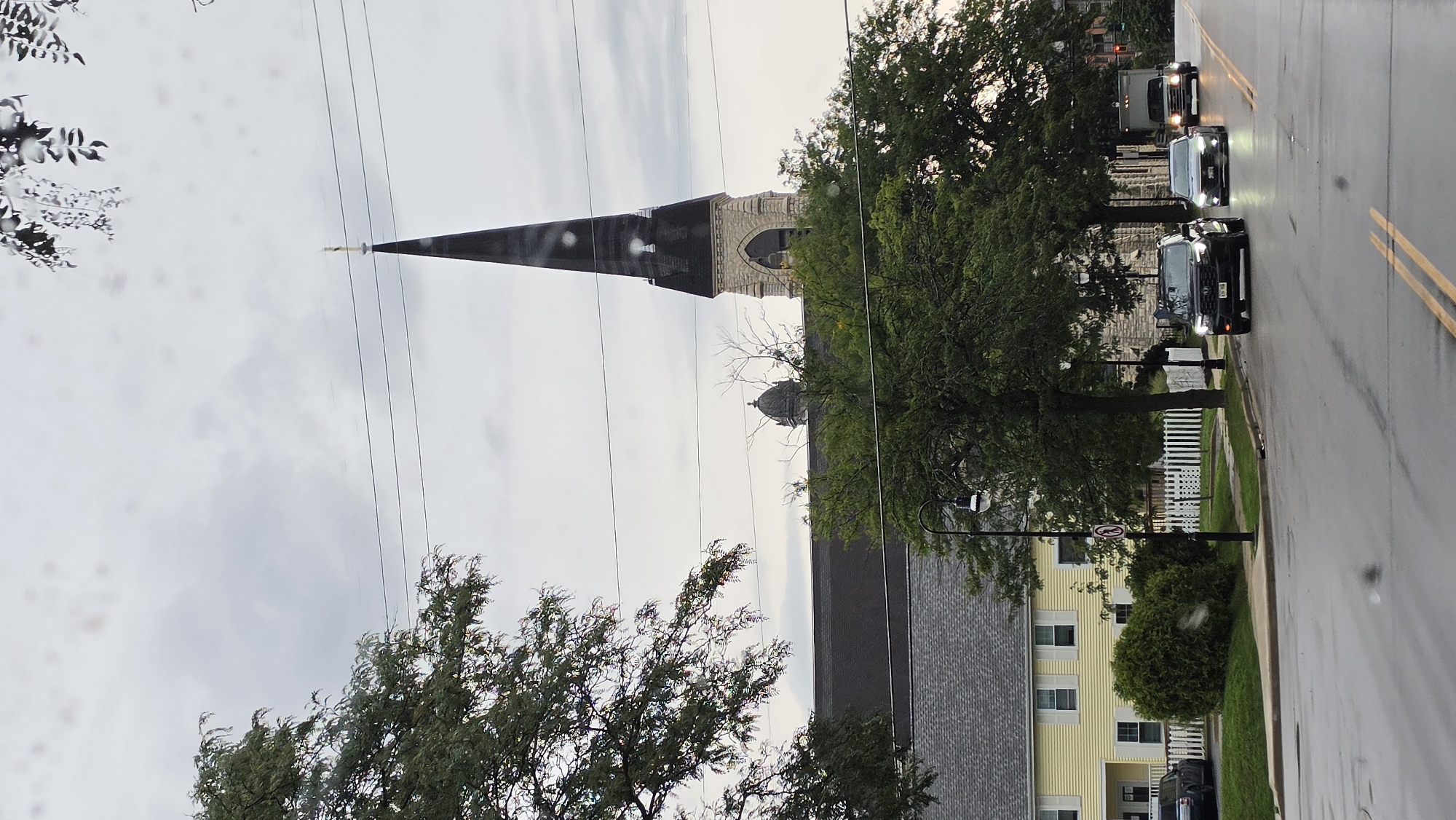 A street view in Waukesha, Wisconsin, shows a dark, overcast sky with some light filtering through. On the left, a large green tree with sparse leaves partially obscures the view. A multi-story yellow building with white trim and shutters stands behind a white picket fence. A black SUV is parked in front of the building. A church steeple, pointed and dark with a cross at its peak, rises above the tree line in the background. The church has stone and dark siding with arched windows near the base of the steeple. A traffic light is visible to the right of the yellow building. The wet asphalt street in the foreground reflects the sky and the headlights of cars. Two cars, a black SUV and a white sedan, are driving towards the viewer. A large white box truck is following them. A black street lamp with a white light fixture is positioned near the sidewalk. There are power lines running horizontally across the frame.