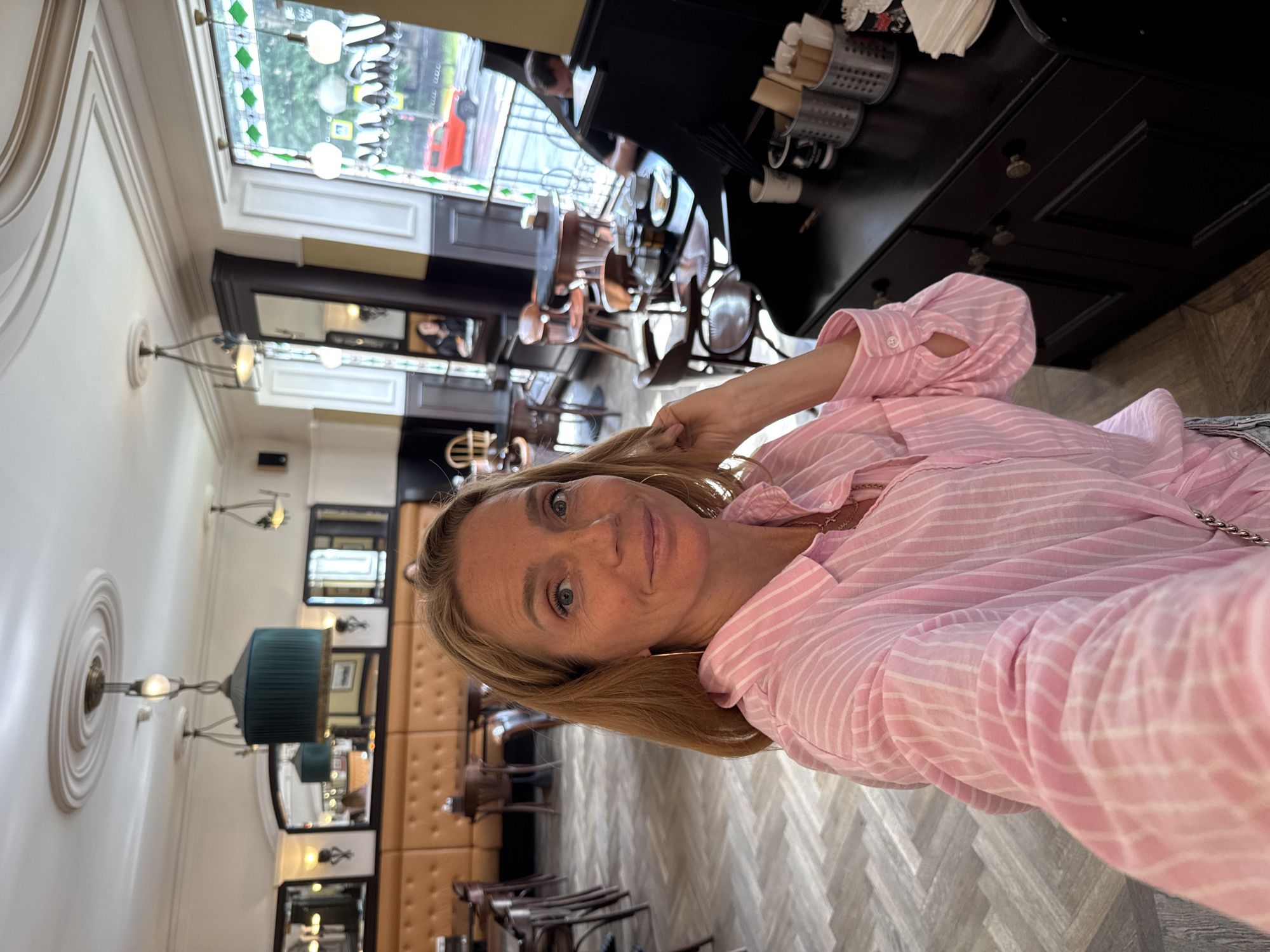 A woman with blonde hair and blue eyes is taking a selfie in what appears to be a cafe or restaurant. She is wearing a pink and white striped button-down shirt and a necklace. She is standing in the foreground, with the background showing tables, chairs, and windows. The cafe has light-colored flooring with a wood-plank pattern and light-colored walls. There are several decorative ceiling elements visible. Through the windows, an exterior scene with trees and a red car is visible. The overall atmosphere is bright and casual. Given the specific architectural details like the ornate ceiling rosettes and the style of the windows, this cafe could be located in Saint Petersburg, Russia.