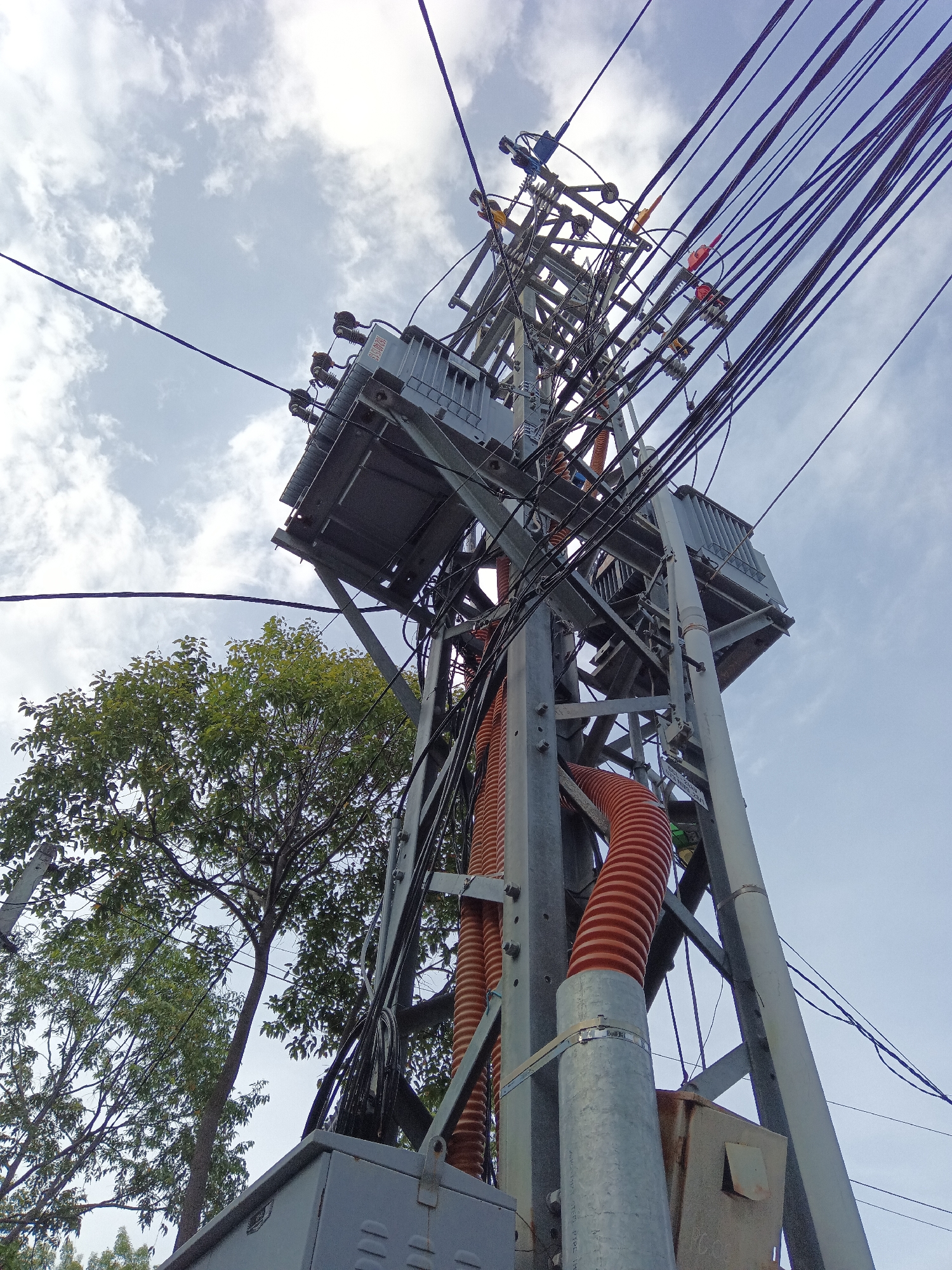 A low-angle shot captures a densely wired electricity pole situated in Nha Trang, Vietnam. The pole, constructed from grey metal beams, is adorned with numerous electrical transformers, insulators, and a chaotic tangle of black wires against a pale blue sky with scattered white clouds. Orange corrugated conduits snake down the pole, carrying cables. To the left, a lush green tree with dense foliage partially obscures the pole, its branches extending towards the structure. The base of the pole is partially visible, with a grey electrical box and some smaller metal boxes. The scene suggests an urban environment where electrical infrastructure is a prominent feature.