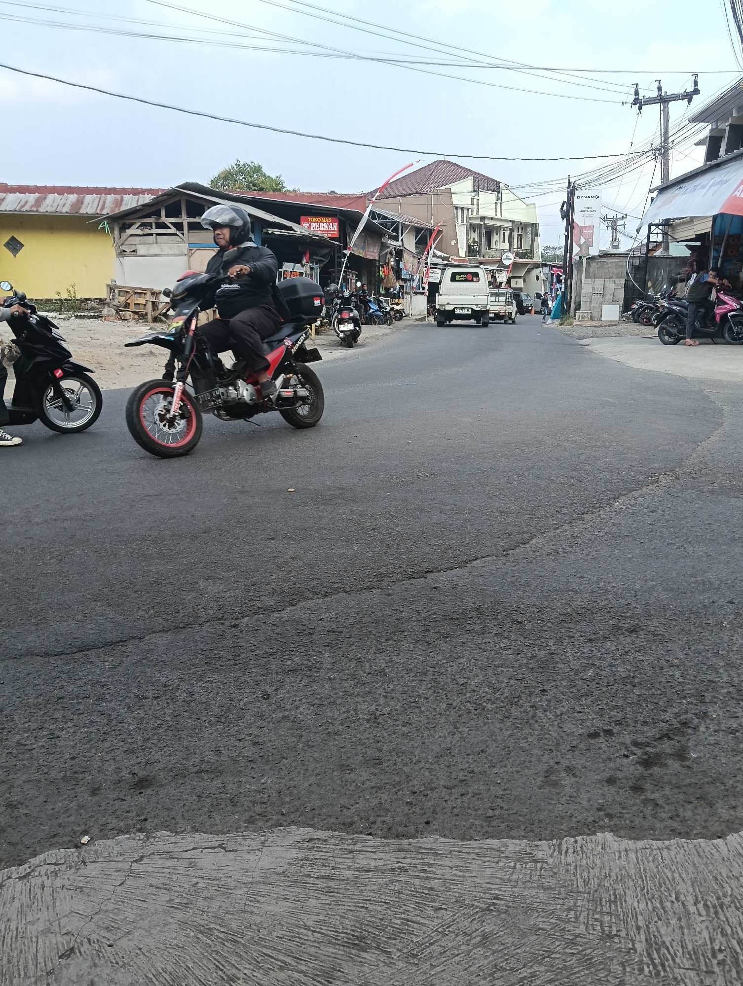 This is an eye-level, medium shot of a street scene in Puspahiang, Indonesia, with a focus on a motorcyclist in the foreground. The road surface is asphalt and appears worn. A man wearing a black helmet and a black jacket is riding a red and black motorcycle. The motorcycle has a black storage box mounted on the rear and a license plate. To the left of the main motorcyclist, another person is partially visible on a black motorcycle. In the background, there are buildings with corrugated metal roofs, some appearing to be shops or small businesses. A white truck is also visible on the road. Overhead, power lines and utility poles crisscross the sky, which is overcast. The overall impression is a busy, everyday street in a semi-urban or rural Indonesian setting.
