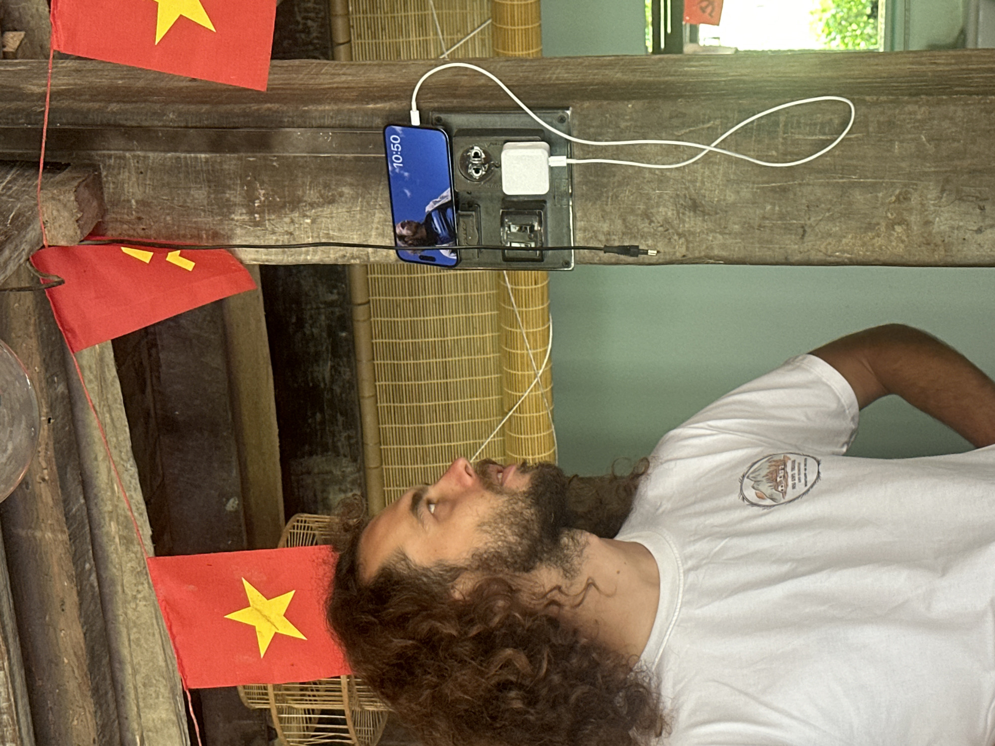 The image captures a man with long, curly brown hair and a full beard, wearing a white t-shirt with a distinct, though largely illegible, graphic logo on the chest. He is positioned looking upwards and slightly to his right, with a neutral or contemplative expression.
Above the man, mounted on a rough-hewn wooden beam, is a black multi-port charging station. A dark-colored smartphone is plugged into the station, displaying "10:50" on its screen and a partial image of a person as its wallpaper. A white charging cable connects the phone to the station, and another white cable leads from a white power adapter also plugged into the station. A black cable with a small jack-like connector is visible near the station.
Several small Vietnamese flags, characterized by their red background and yellow star, are strung along the wooden beams in the foreground and mid-ground, serving as prominent cultural identifiers.
In the background, woven bamboo blinds or screens provide a textured backdrop, suggesting a traditional or rustic architectural style. A light green wall is visible behind the blinds, and a window shows blurred greenery, indicating daylight and an outdoor view.
The scene suggests a late morning setting, given the time displayed on the phone. The presence of Vietnamese flags and rustic wooden elements, combined with modern electronics, points to a location within Vietnam, such as Du Già Commune, where traditional settings often incorporate contemporary conveniences.