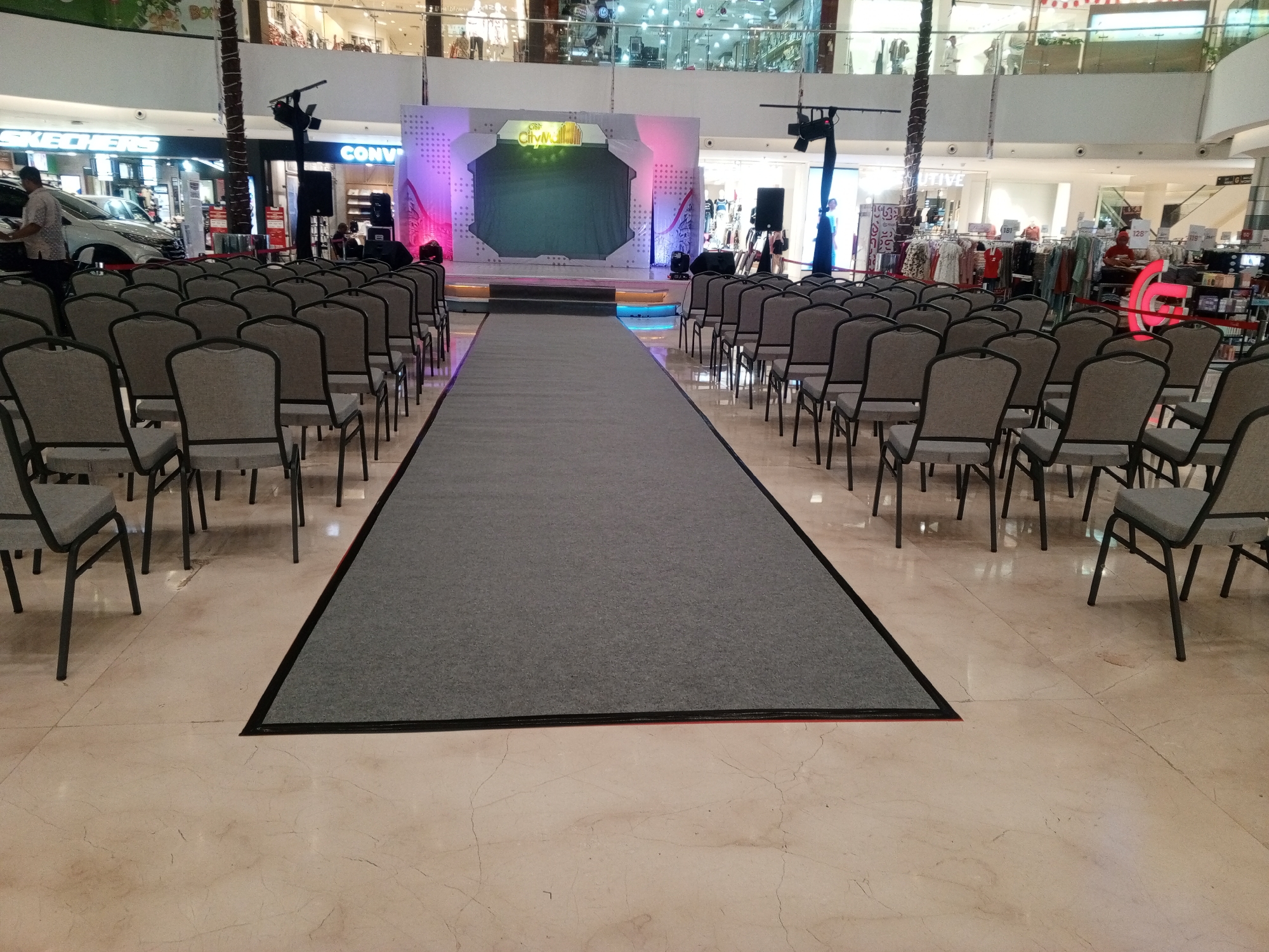 The image depicts a fashion show or event setup within a mall. A gray carpeted runway extends from the foreground towards a stage with a large screen and backdrop featuring the "City Walk" logo. Rows of gray chairs are arranged on either side of the runway. In the background, mall storefronts are visible, including "Skechers" and "Converse," indicating the location is likely within a mall in Cibinong, Indonesia, given the presence of these brands and the architectural style. A few individuals are present in the scene, including a person standing near a white car on the left and a person in an orange shirt on the right, possibly staff or attendees. The lighting suggests an indoor environment with some artificial stage lighting.