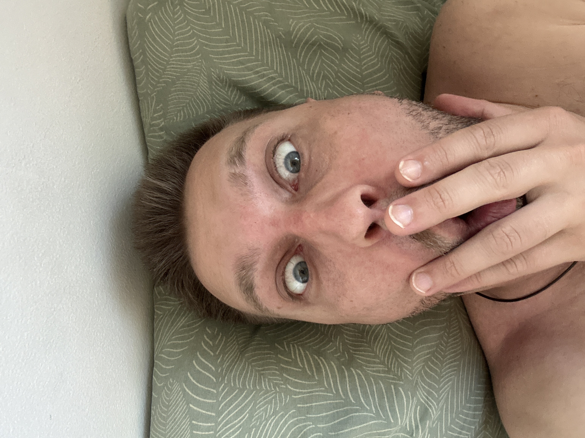 A close-up, high-angle, selfie-style photograph shows a person's face, with their eyes wide open and their hand covering their mouth, appearing surprised or shocked. The person has light skin, brown hair, and blue eyes. They are lying down on a light green pillow with a white leaf pattern, and a white wall is visible above the pillow. The person is wearing a black cord necklace. The scene is indoors, likely in a bedroom. The location is Zvezdara Urban Municipality, Serbia.