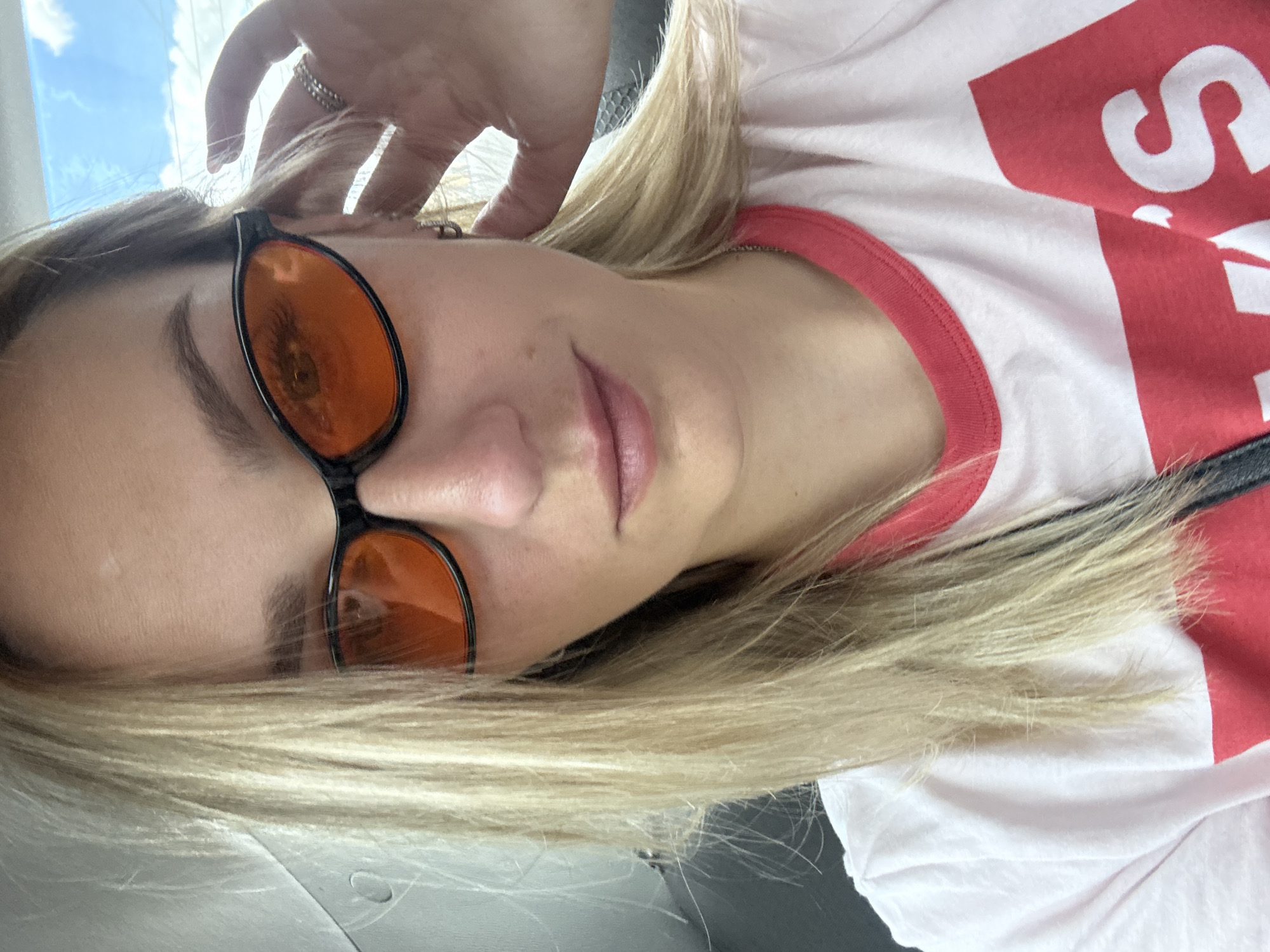 A young woman with blonde hair and sunglasses is taking a selfie in a car. She is wearing a white t-shirt with red lettering. Her left hand is touching her hair near her ear. The interior of the car is dimly lit, and the sky is visible through the window.