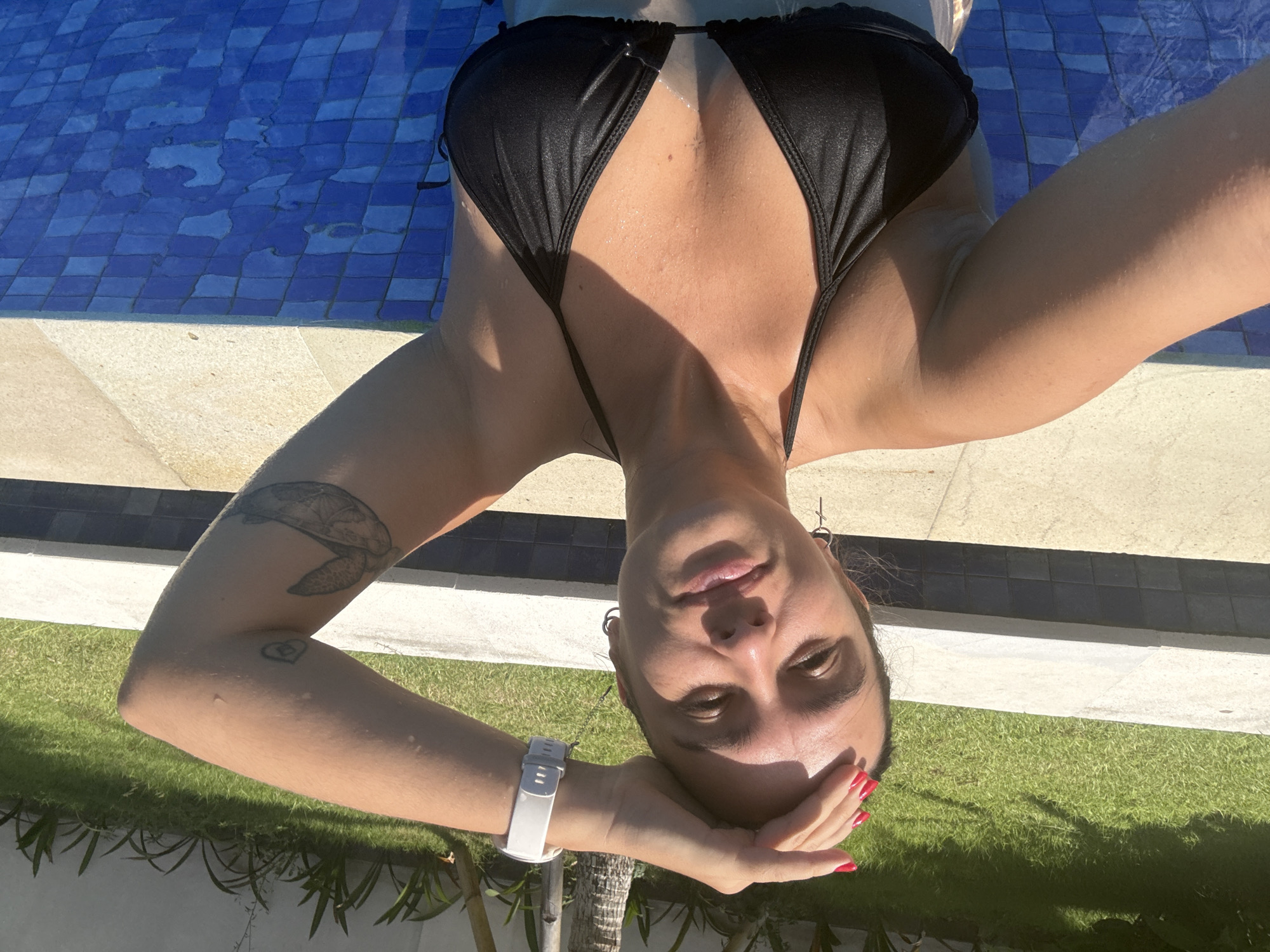 A female individual is depicted in a swimming pool located in North Kuta, Indonesia. The subject is shown from the chest up, with her body partially submerged in the blue-tiled water. She is wearing a black halter-neck bikini top. Her left hand, adorned with red nail polish, is positioned on her forehead. A white smartwatch is visible on her right wrist. She has a silver hoop earring in her left ear and a silver cross-shaped earring in her right ear. Two tattoos are present on her right arm: a small heart-like design on her upper arm and a larger, detailed sea turtle on her forearm. The pool's interior wall consists of blue square tiles, bordered by a light-colored coping. In the background, a well-maintained green lawn with tropical foliage is visible, illuminated by bright daylight.