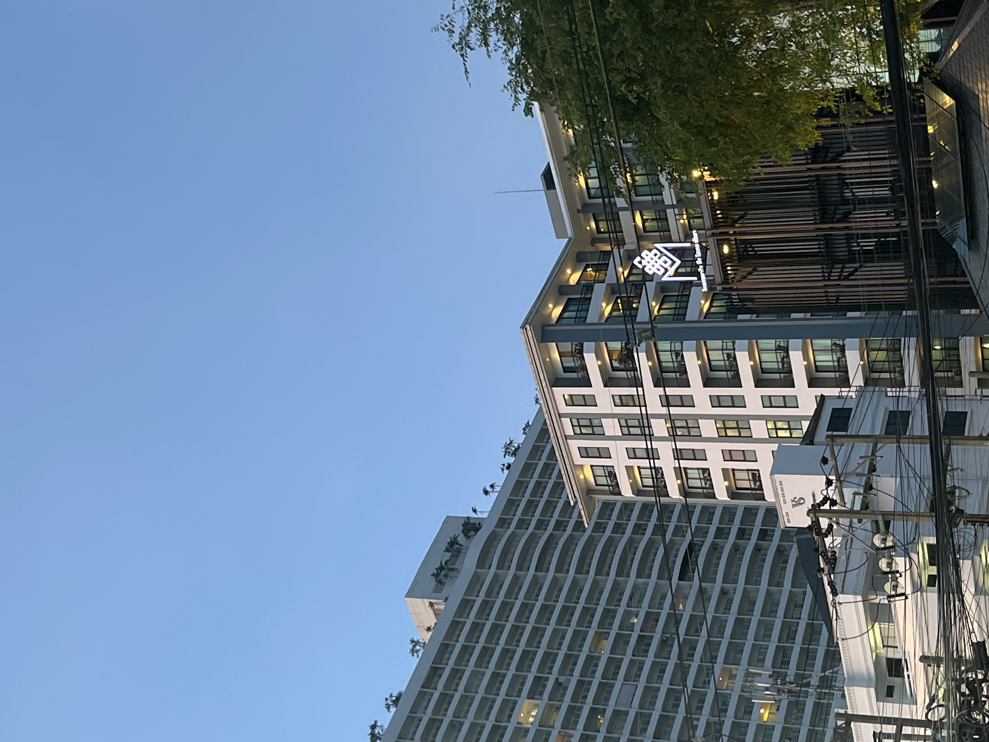 The image depicts a low-angle view of buildings in Pattaya City, Thailand, under a clear blue sky. Two distinct architectural structures are visible. The building to the right is a multi-story structure with a geometric facade featuring grey and white elements, multiple windows, and a well-lit rooftop logo or sign. Adjacent to it is a taller building with a unique wavy or curved pattern across its exterior, also consisting of numerous windows. Electrical poles with a dense network of wires are prominent in the foreground on the right side, partially obscuring the buildings. Trees and greenery are visible at the top right, bordering the building edge. The light suggests it is either early morning or late afternoon.