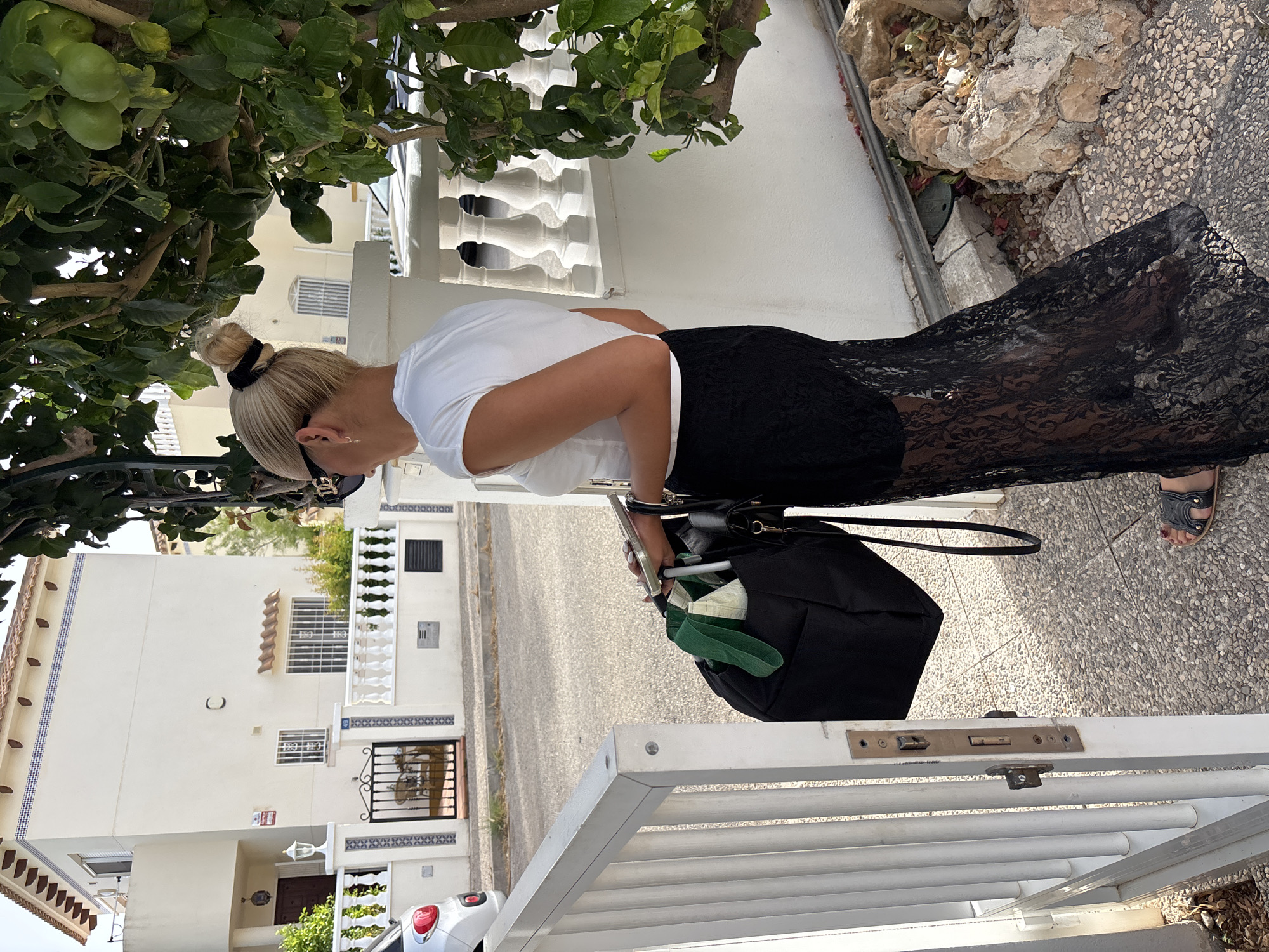 An adult blonde female wearing sunglasses, a white top, and a long black lace skirt is standing near a white gate. She is holding a smartphone and a black bag with green items protruding from it. The background shows a white building with red-tiled roofs, a white fence, and a tree with green leaves. The ground is paved with small, light-colored stones. The scene appears to be a residential street in Orihuela, Spain, characterized by the architectural style of the building and fencing.