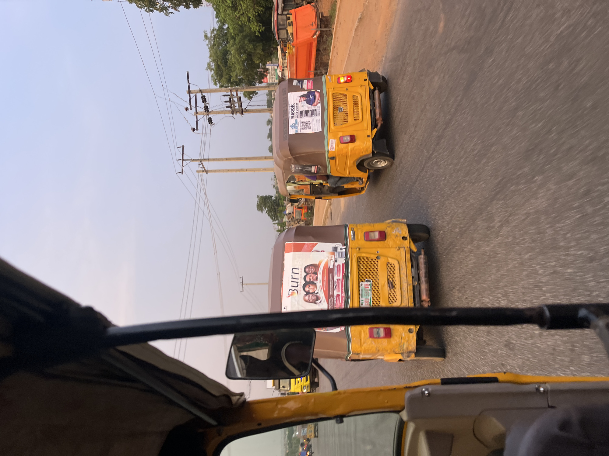 The image, taken from inside a yellow and brown-roofed tuk-tuk, shows two similar tuk-tuks moving along an asphalt road. The tuk-tuk directly in front has a large advertisement on its rear with the word "Burn" visible and images of four people. A license plate is partially visible on the back of the tuk-tuk. The second tuk-tuk has an advertisement with a person's image and text related to "N500K Land Owner". Utility poles are seen along the side of the road, with trees and other vehicles visible in the background. The sky is a light blue, suggesting daytime. The scene is located in Galadimawa Village, Nigeria, based on provided information.