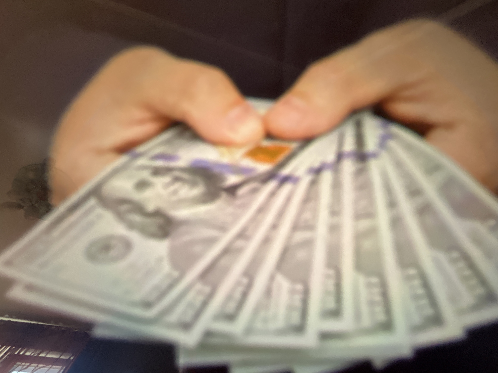 The media file is a close-up, blurred image of a person's hands holding a fan of US dollar bills. The hands are in the center of the frame, and the bills are spread out, showing portions of Benjamin Franklin's portrait on what appear to be $100 denominations. The lighting is dim, suggesting an indoor setting, possibly at night. There are no discernible details about the weather or specific location cues within Gelendzhik, Russia, beyond what can be inferred from the general context. The focus is entirely on the money and the hands, with the background being dark and indistinct. The image conveys a sense of wealth or financial transaction, but without further context, the emotions or specific event remain ambiguous. No visible text is clearly legible on the currency due to the blurriness and the angle.