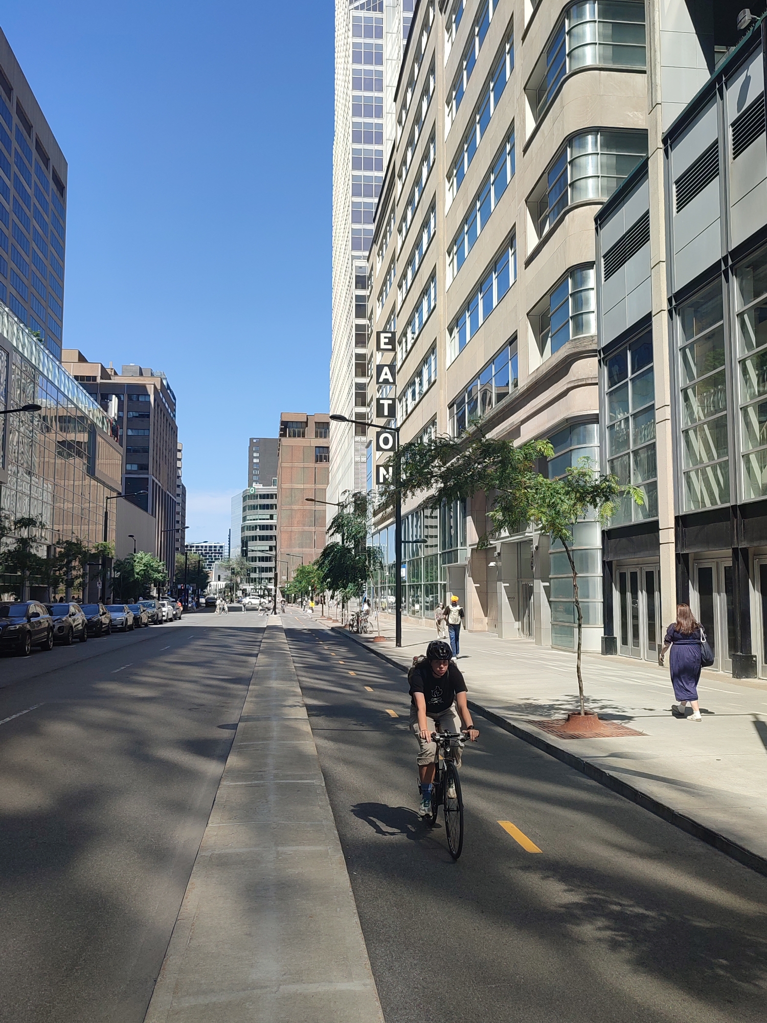 The image depicts a street scene in Montreal, Canada, on a clear, sunny day. The street is lined with tall, modern buildings on both sides, with a clear blue sky overhead. A cyclist wearing a black t-shirt, beige shorts, and a black helmet is riding a bicycle down the street, which has a dedicated bike lane marked with yellow lines. To the right of the cyclist, a woman in a dark blue dress and white shoes is walking on the sidewalk. Other pedestrians are visible further down the street, and parked cars line the left side of the road. The buildings feature large windows, and one prominent building on the right has "EATON" spelled vertically on its facade. Trees are planted along the sidewalk, casting shadows on the street.