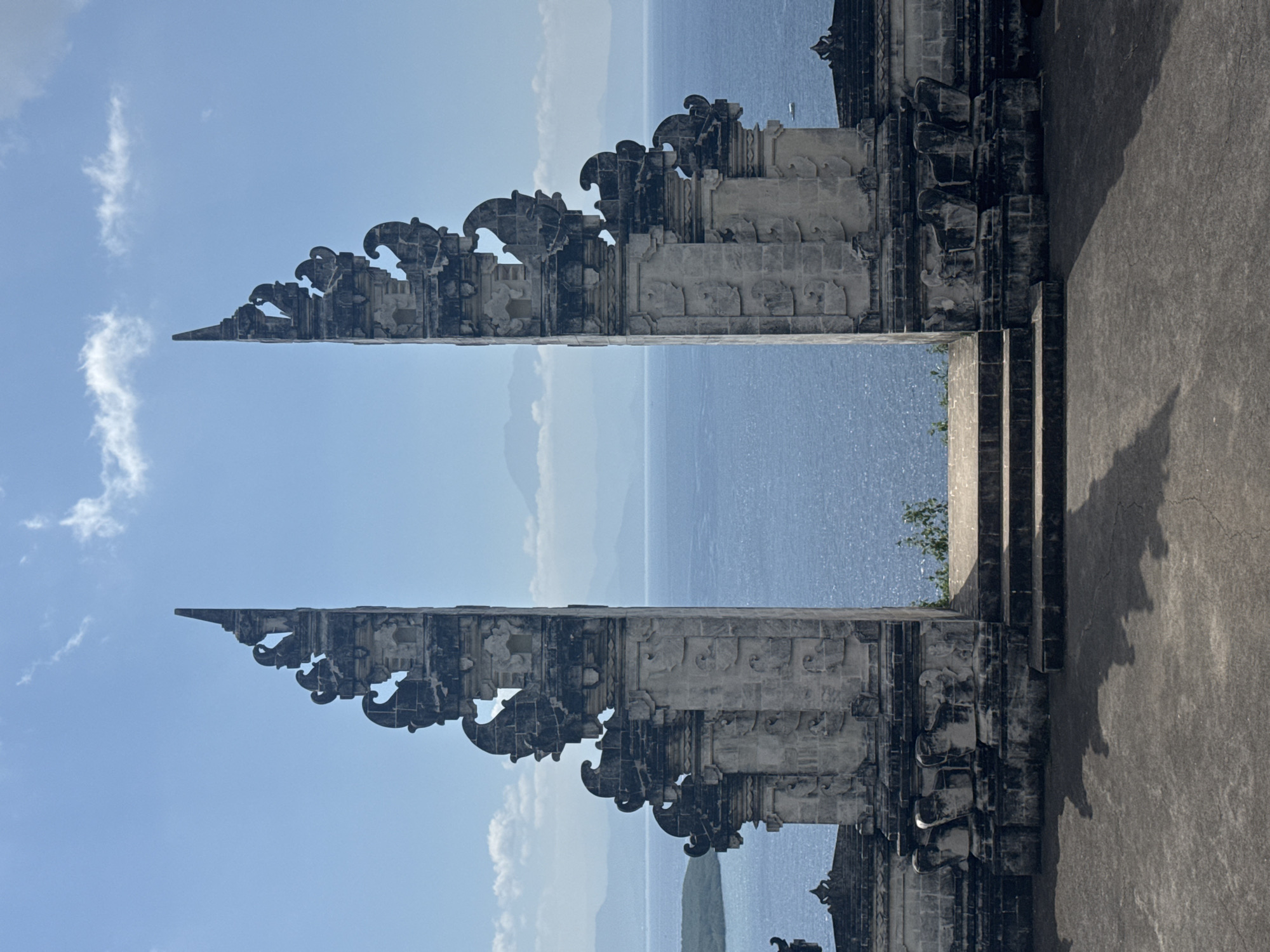 The image displays a symmetrical composition featuring two tall, ornate, grey stone gate pillars, characteristic of a Balinese Candi Bentar, often referred to as the "Gates of Heaven." These structures are intricately carved with traditional Hindu motifs, exhibiting a weathered texture. They stand prominently on a flat, paved foreground, where their distinct shadows are cast, indicating strong sunlight from above and slightly behind the viewer. A set of three stone steps leads up to the opening between the gates.
Beyond the gates, a vast expanse of sparkling blue ocean stretches towards the horizon, where distant, hazy, blue-tinted landmasses or mountains are visible. The sky above is a clear light blue with a few scattered, wispy white clouds. A small, light-colored boat or vessel is discernible on the ocean surface to the right of the right gate. Sparse green foliage is present at the base of the gates, just before the apparent drop-off towards the sea. No human subjects are visible within the frame.
The location depicted is Pura Lempuyang Luhur, a temple situated in East Bali, Indonesia.