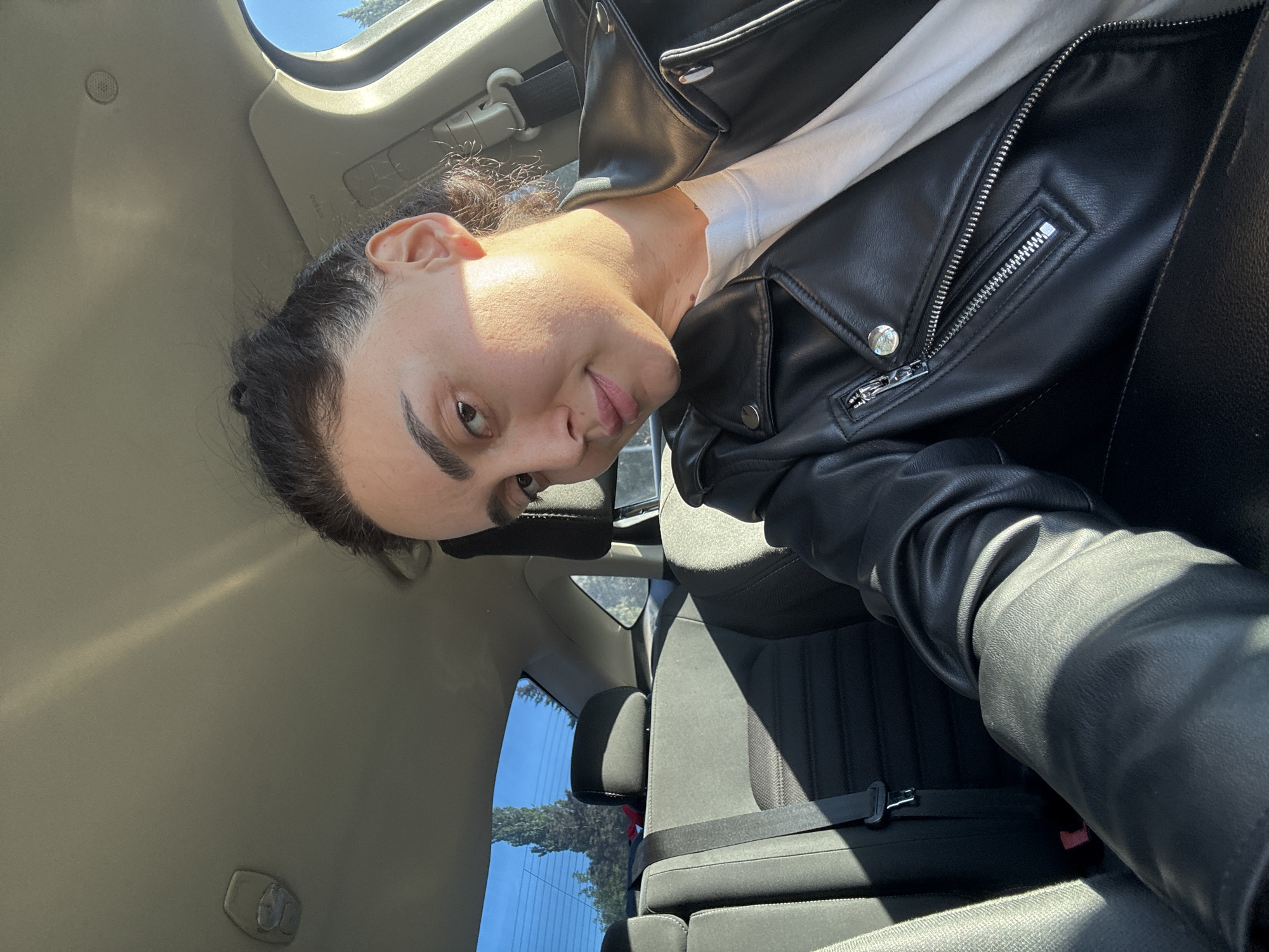 The image shows a person in the front seat of a car. The person has dark hair pulled back and is wearing a white shirt and a black leather jacket. The car's interior is visible, including the seat, seatbelt, and ceiling. Outside the car, trees and a blue sky can be seen. The location of Tbilisi, Georgia is not verifiable from the information provided in the image.