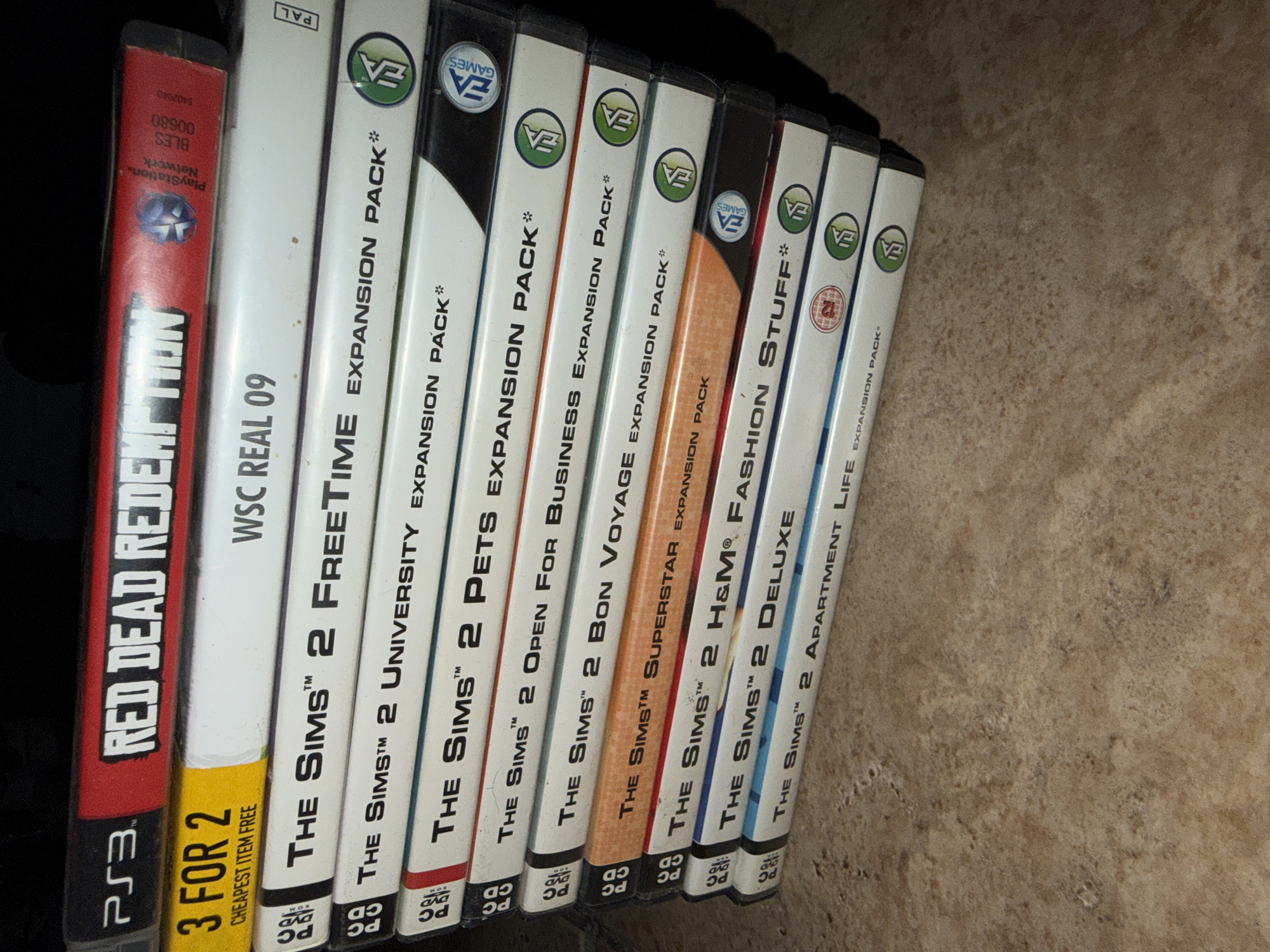 The image shows a stack of video game cases. The top case is for "RED DEAD REDEMPTION" for PS3. Below that is a yellow case with "3 FOR 2 CHEAPEST ITEM FREE" printed on it, and "WSC REAL 09" to the right. The remaining cases are for various "The Sims 2" expansion packs, including "FreeTime", "University", "Pets", "Open For Business", "Bon Voyage", "Superstar", "H&M Fashion Stuff", "Deluxe", and "Apartment Life". Several of the "The Sims 2" cases have the EA Games logo. The background is dark, and the games are resting on a textured surface that appears to be a countertop or floor. The image does not provide any information about the location in Sharm El Sheikh, Egypt.