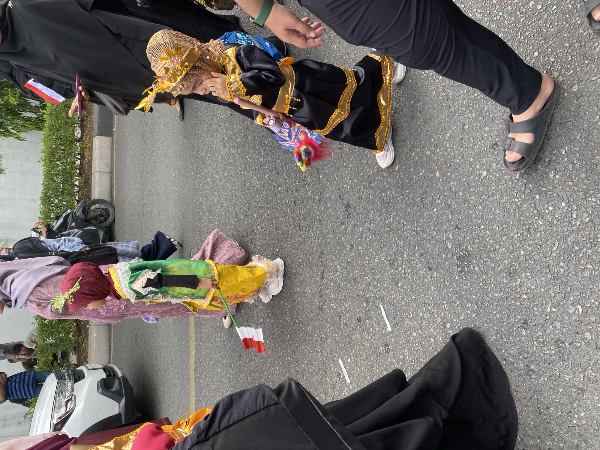 In Banda Aceh, Indonesia, a procession is underway on a paved street. Two young girls are the central figures. The first girl, on the left, is wearing a red hijab and a green and yellow traditional outfit, holding a small Indonesian flag. The second girl, on the right, is adorned in a black and gold outfit with a golden crown, appearing to touch her face. Several adults are also present, mostly wearing black Islamic clothing, some holding phones or personal items. The background shows a white car, a motorcycle, and lush green bushes, indicating an urban setting. The ground is littered with small white papers.