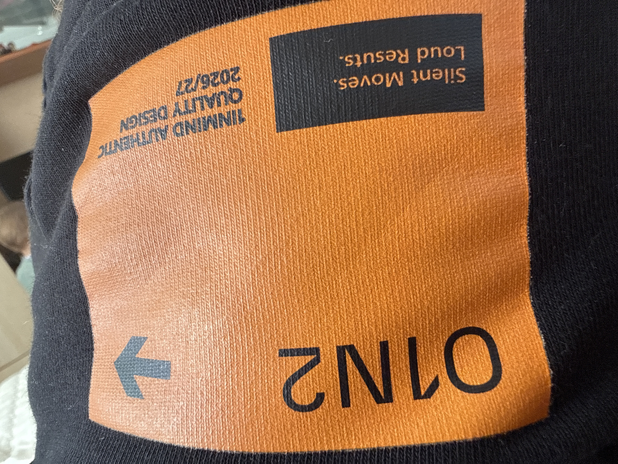 The image shows a close-up of a black t-shirt with an orange rectangular graphic printed on it. The graphic contains text and symbols. Visible text includes "INMIND AUTHENTIC QUALITY DESIGN 2026/27" printed vertically on the left side of the graphic. To the right of this, on a black background within the orange rectangle, is the text "Silent Moves. Loud Results.". On the lower right portion of the orange graphic, large black numbers read "1N12 01N10". A dark grey arrow pointing left is also visible on the lower left side of the orange graphic. The t-shirt appears to be made of a textured fabric, possibly cotton. The background is out of focus but suggests an indoor setting. The lighting appears to be artificial.