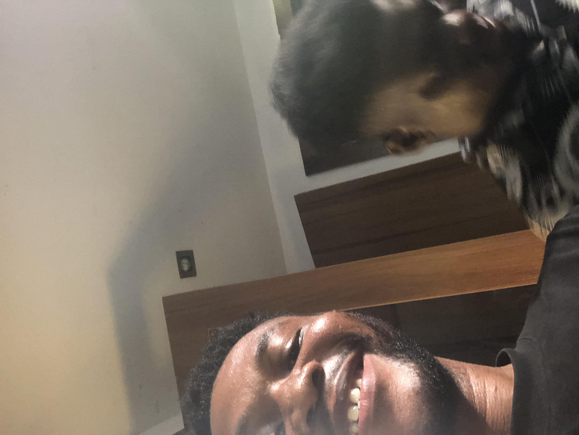 The image captures a close-up, angled view of two individuals in an indoor setting. In the foreground, a person with dark skin and a beard is prominently featured, smiling broadly with white teeth visible. Their face is well-lit, showing a shiny or sweaty sheen, and they appear to be looking upwards.
Behind the foreground subject, partially obscured, is another person leaning over. Only the top of their head, dark hair, and a portion of their face looking downwards are visible. They are wearing a dark, possibly patterned, garment.
A light-colored wall serves as the background, with a horizontal wooden beam or shelf structure visible between the two individuals. A small, dark-framed object, possibly a picture or light switch, is affixed to the wall in the upper left corner of the frame. The overall impression is one of a cheerful and intimate interaction, with the foreground subject's wide smile conveying happiness or amusement. The scene appears to be well-lit, suggesting an indoor daytime setting or a brightly illuminated room. There is no visible text in the image.