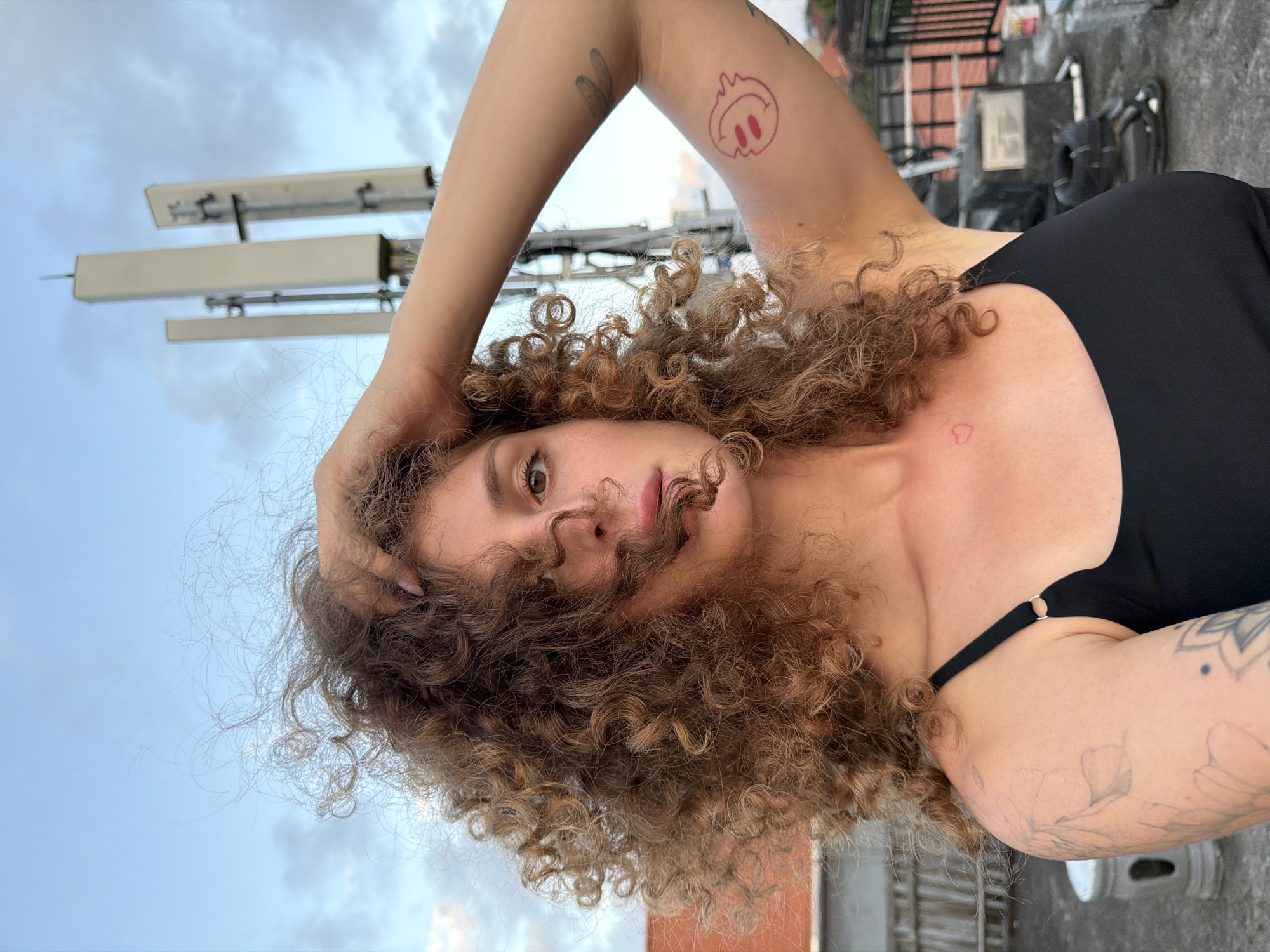 A medium shot captures a woman with voluminous curly brown hair touching her head, looking towards the camera. She is wearing a black tank top. Her left arm is raised and bent at the elbow. A red tattoo of a smiling face is visible on her upper left arm. Another tattoo of a heart is on her upper chest. The background features a cell tower against a cloudy sky, and a rooftop with some equipment. The image was taken in North Kuta, Indonesia.