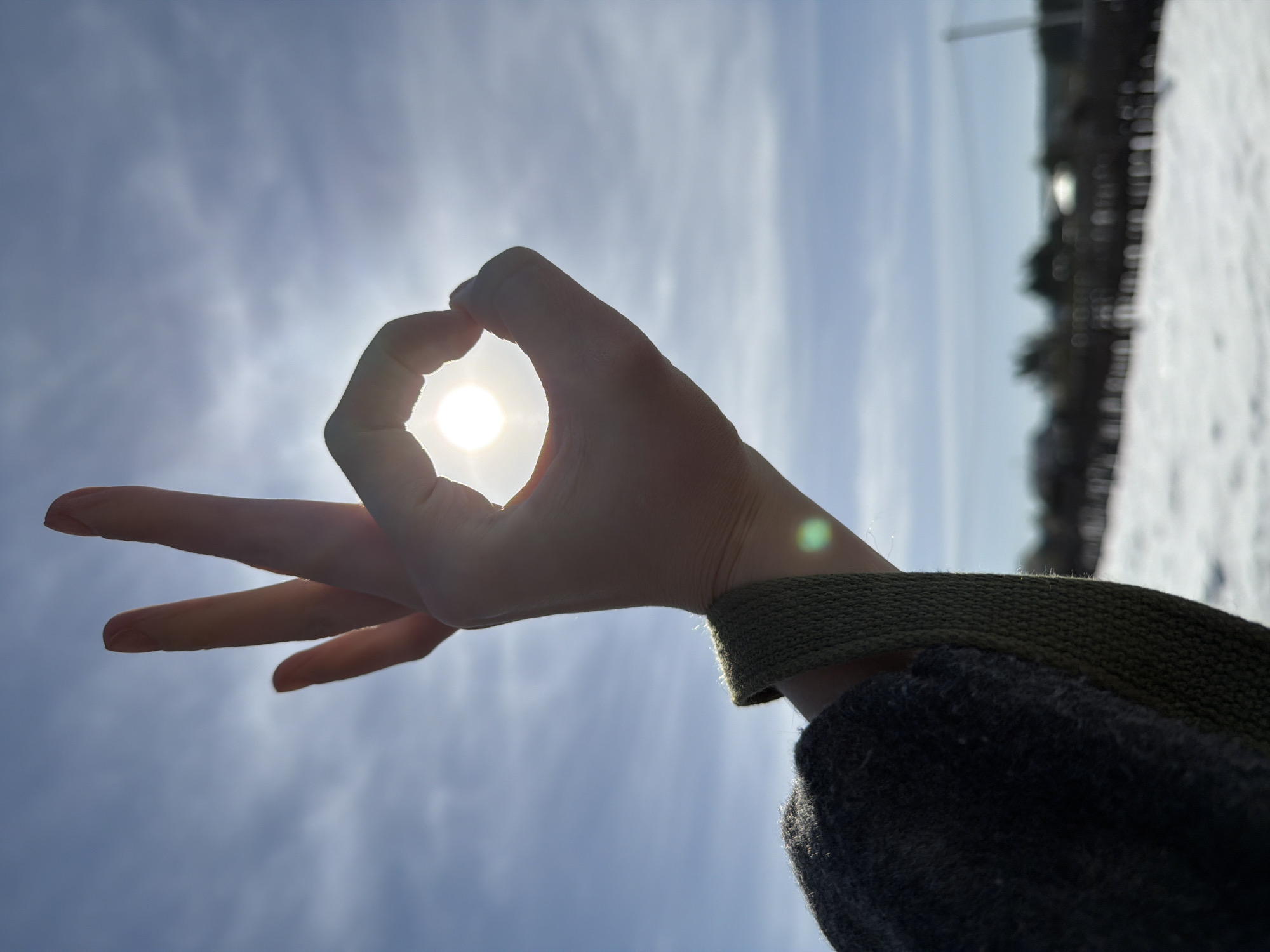 A hand is held up against a bright sky, with the sun shining through the "OK" gesture formed by the thumb and forefinger. The middle, ring, and pinky fingers are extended outwards. The hand is attached to an arm clad in a dark sweater with a textured, dark green band around the wrist. The background features a bright, slightly clouded sky. The horizon line reveals snow-covered ground and blurred dark shapes that suggest distant trees or buildings. This photo appears to be taken in daylight outdoors. While the image's location is not explicitly stated, the prompt does suggest that this image may have been taken in the city of Chaikovsky, Russia.