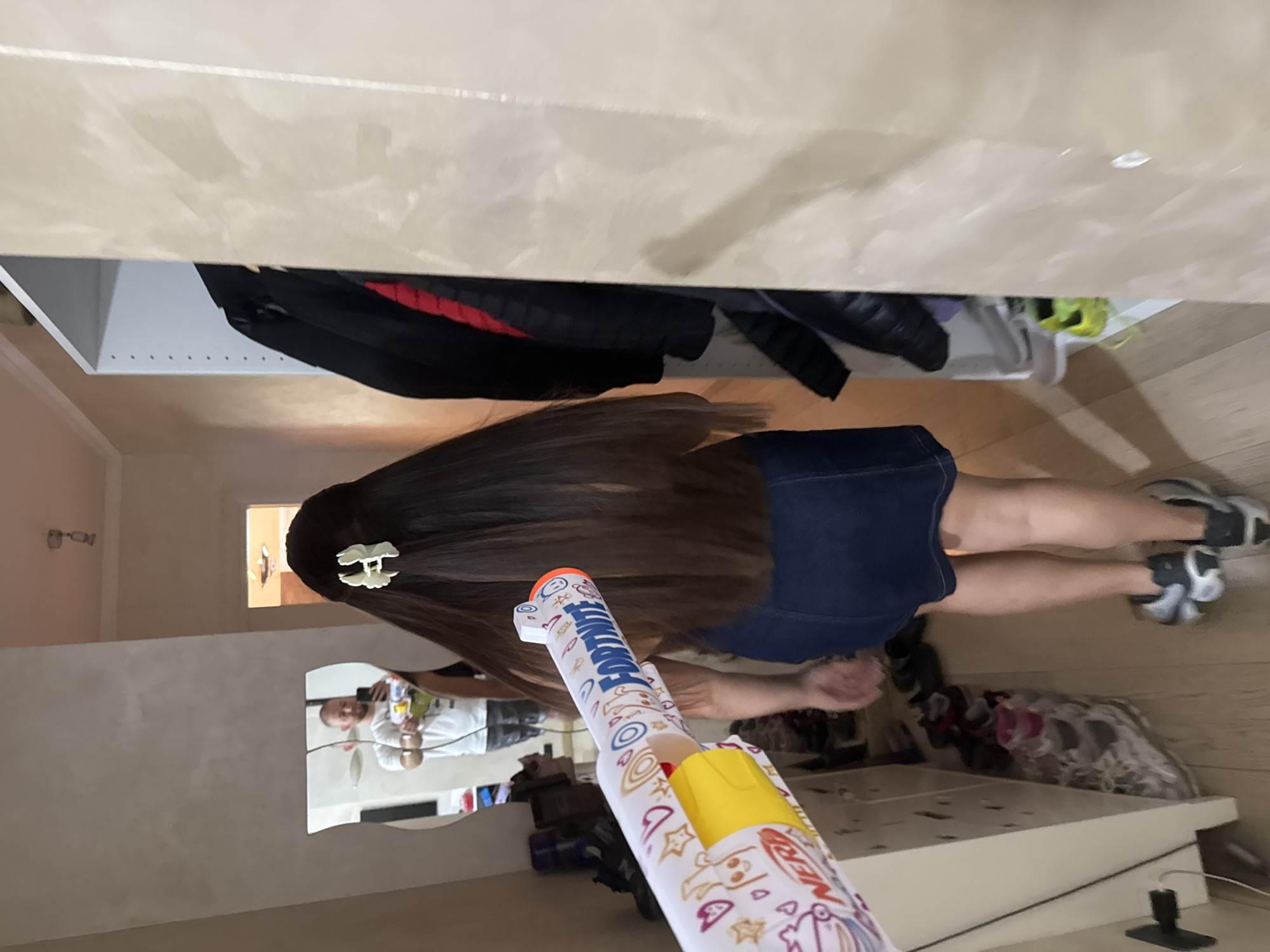 The image shows a person holding a Nerf Fortnite dart blaster in a room. The person is wearing a dark blue skirt and gray running shoes. Their long brown hair is adorned with a white hair clip. The room appears to be a bedroom with a wardrobe or closet visible in the background, containing various clothing items. A mirror reflects a man holding a phone, likely taking the picture. The environment suggests a private indoor space, possibly a residence. There is no indication of the city of Rome, Italy.