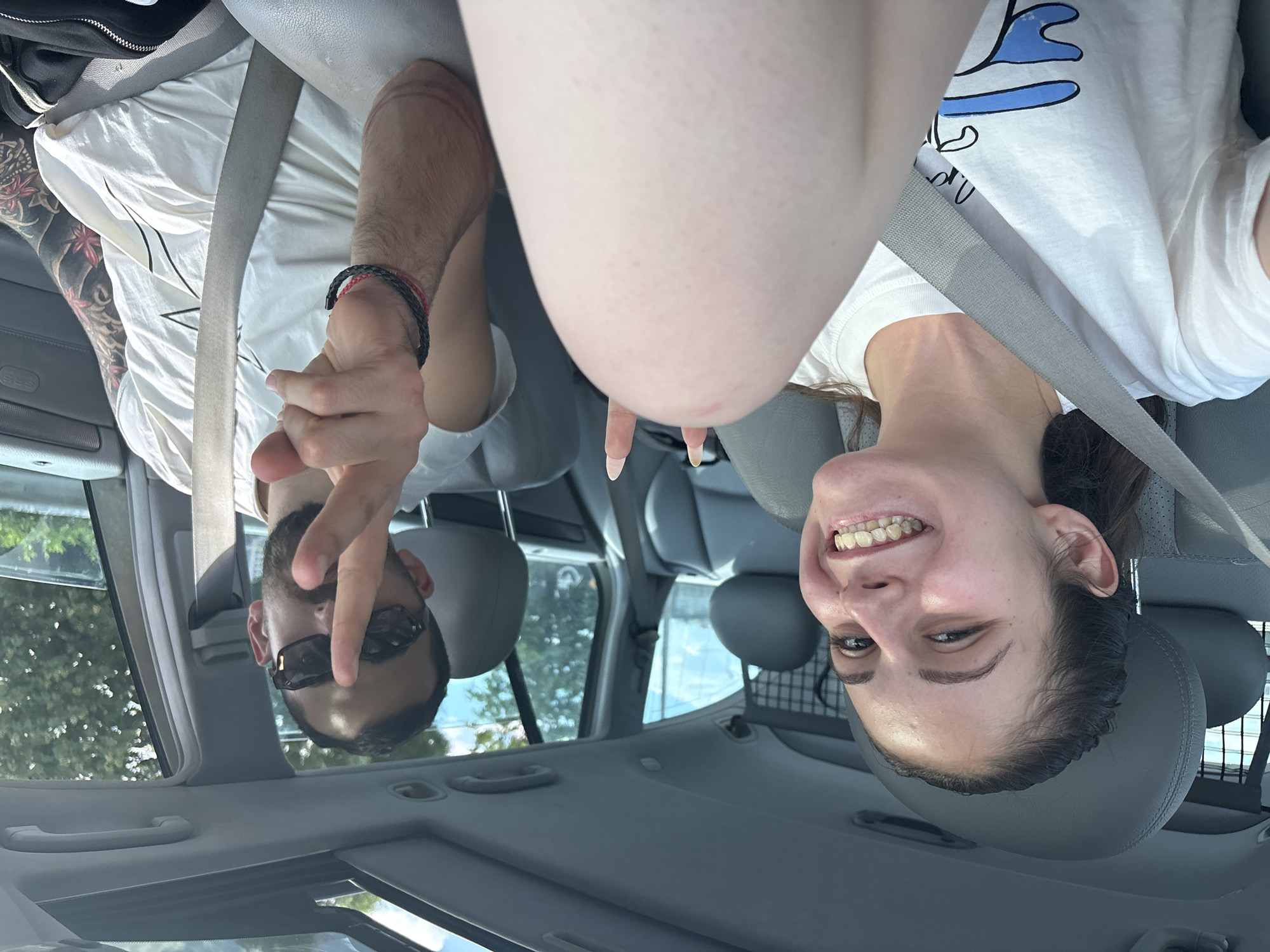 The image shows two individuals, a man and a woman, inside a car. The image is oriented upside down. The woman, on the right side of the frame, is smiling and wearing a white t-shirt with a graphic. She is wearing a seatbelt. The man, on the left side of the frame, is wearing sunglasses, a white t-shirt, and has tattoos on his arm. He is making a peace sign with his fingers. The interior of the car is visible, including the seats, seatbelts, and dashboard. Outside the car, trees and a glimpse of a metal fence or gate can be seen. The city and country are not discernible from the image.