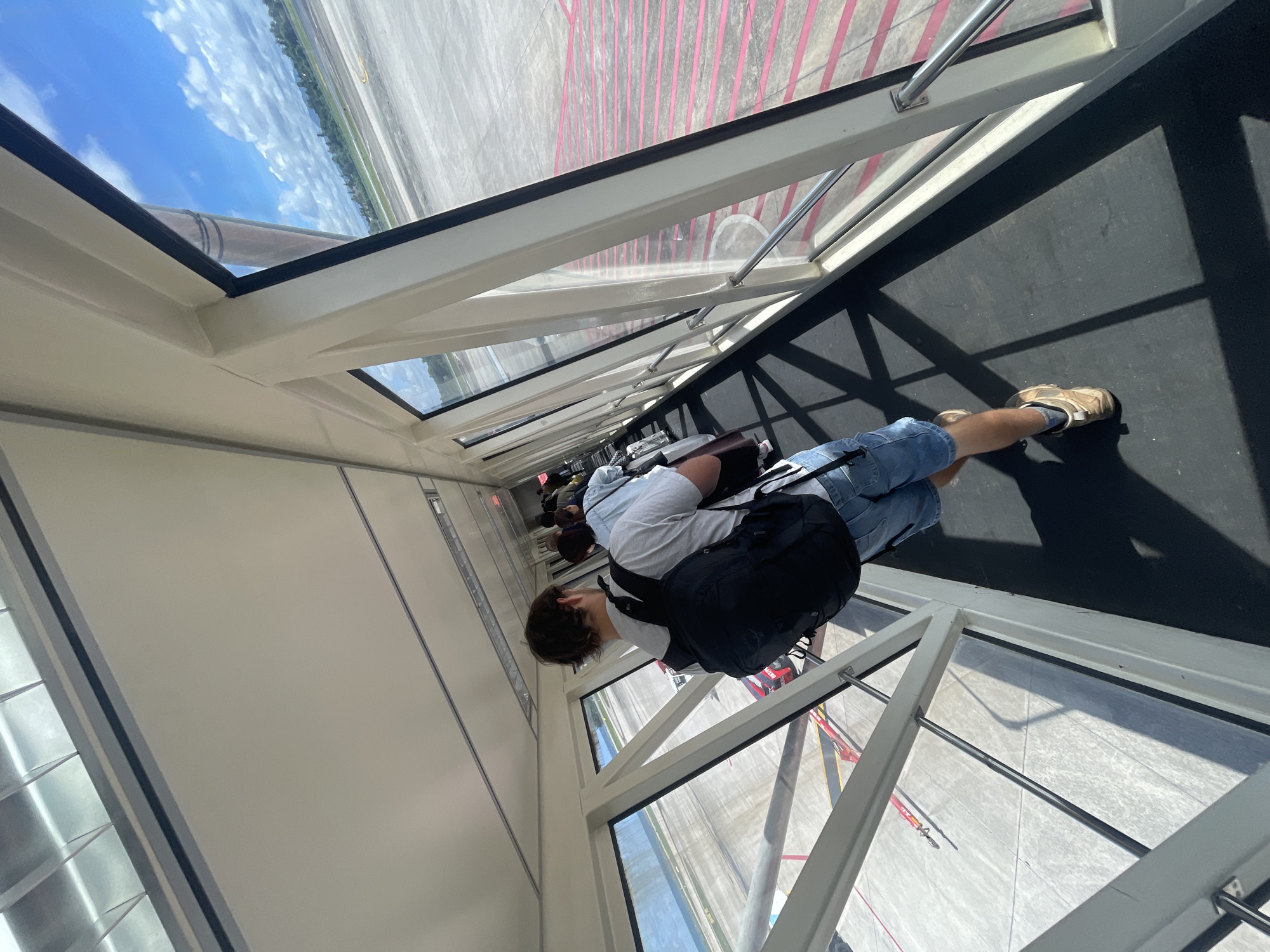 The image shows a group of people walking through an airport jet bridge towards an airplane. The perspective is from behind one of the travelers, a male with a black backpack and denim shorts, facing forward. The jet bridge has large glass windows on the right side, offering a view of the tarmac, runway, and a cloudy blue sky. Red lines are painted on the tarmac. The environment appears to be an airport located in Mai Khao, Thailand, suggested by the tropical landscape visible in the distance. The people are queuing and moving in a single file line, implying they are boarding the aircraft.