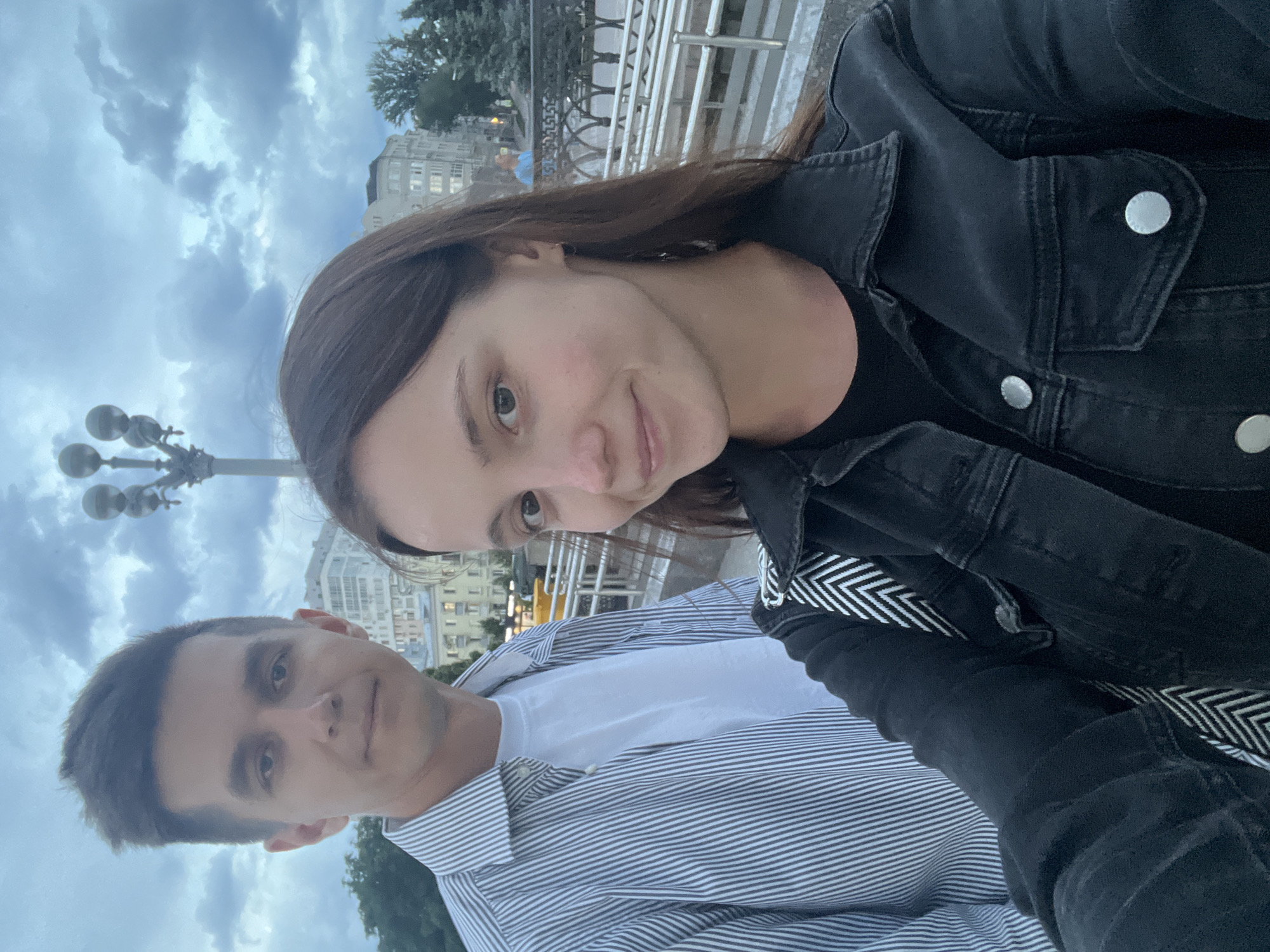 A selfie is taken outdoors in an urban environment under a cloudy sky. In the foreground, a young woman with brown hair smiles towards the camera, wearing a black denim jacket over a black t-shirt. To her left and slightly behind her, a young man with short, dark hair is visible, wearing a striped collared shirt over a white t-shirt. The background features buildings and trees, suggesting a park or public space. The railing of a bridge or elevated walkway is visible between the couple and the background. The overall lighting is dim, consistent with overcast weather. The location is specified as Novokuybyshevsk, Russia.