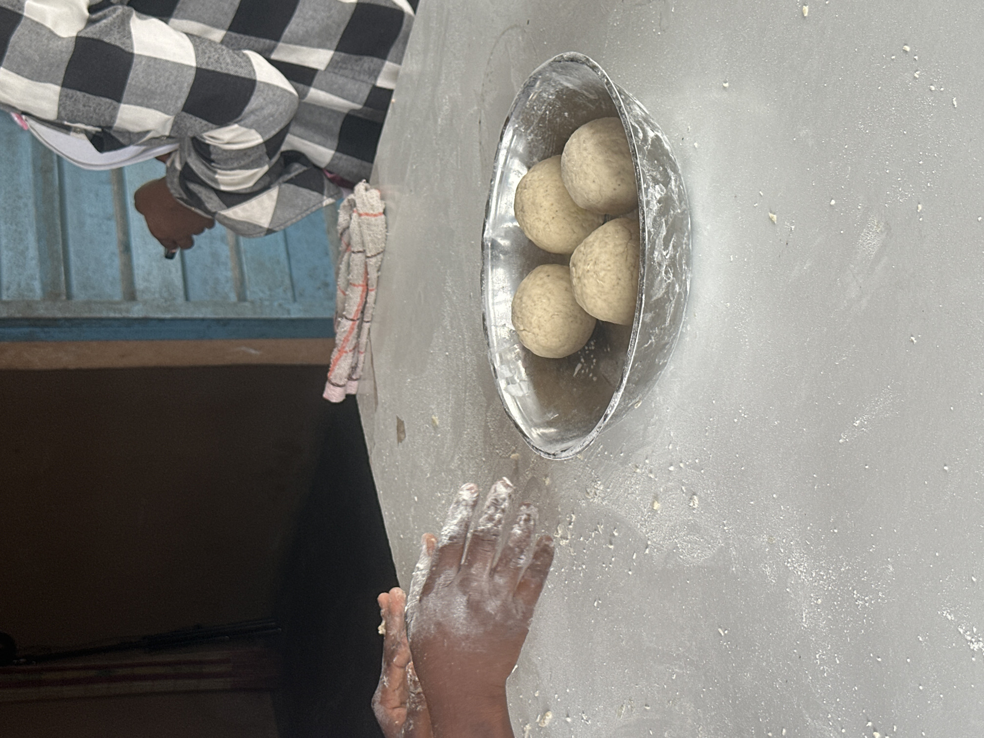 In a scene suggestive of food preparation, a person's hands, dusted with white flour, are actively engaged in shaping dough. The hands are positioned over a surface also lightly coated with flour. To the right of the hands, a metal bowl contains four round portions of dough. In the background, a person wearing a black and white checkered garment is visible, holding what appears to be a small object, possibly a pen or stylus, near a blue louvered window. A folded cloth with red and white stripes lies on the table between the hands and the person in the checkered garment. The overall environment appears to be indoors, with a dark background suggesting a wall or interior space. The presence of flour and dough indicates a cooking or baking activity, likely taking place in a domestic setting in Badagry, Nigeria.